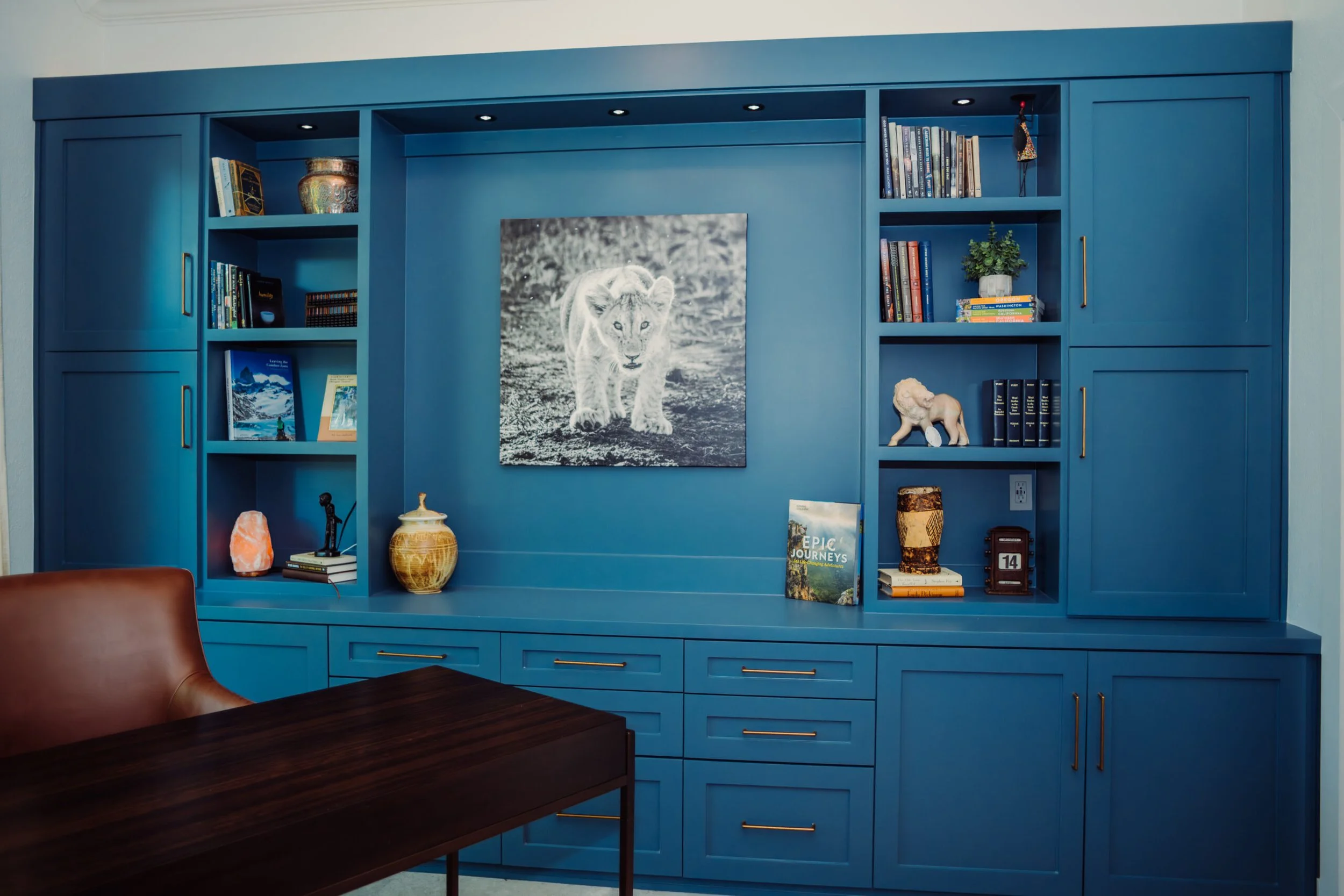 A blue built-in wall unit with shelves filled with books, decorative items, and art pieces in a room with a brown leather chair and a dark wooden table.
