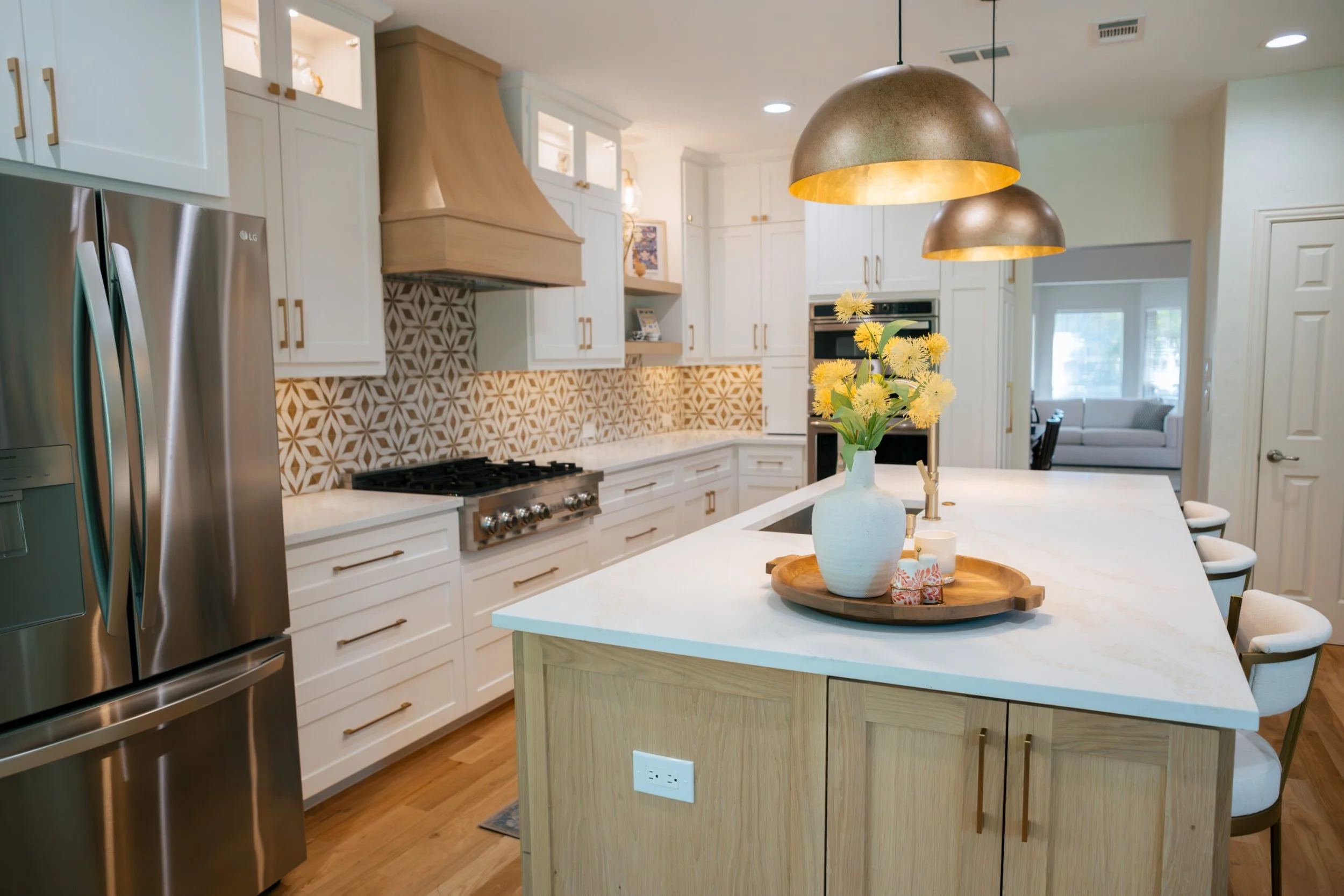 Modern kitchen with white cabinets, wooden accents, gold hardware, stainless steel refrigerator, and a central island with a vase of yellow flowers and a wooden tray.