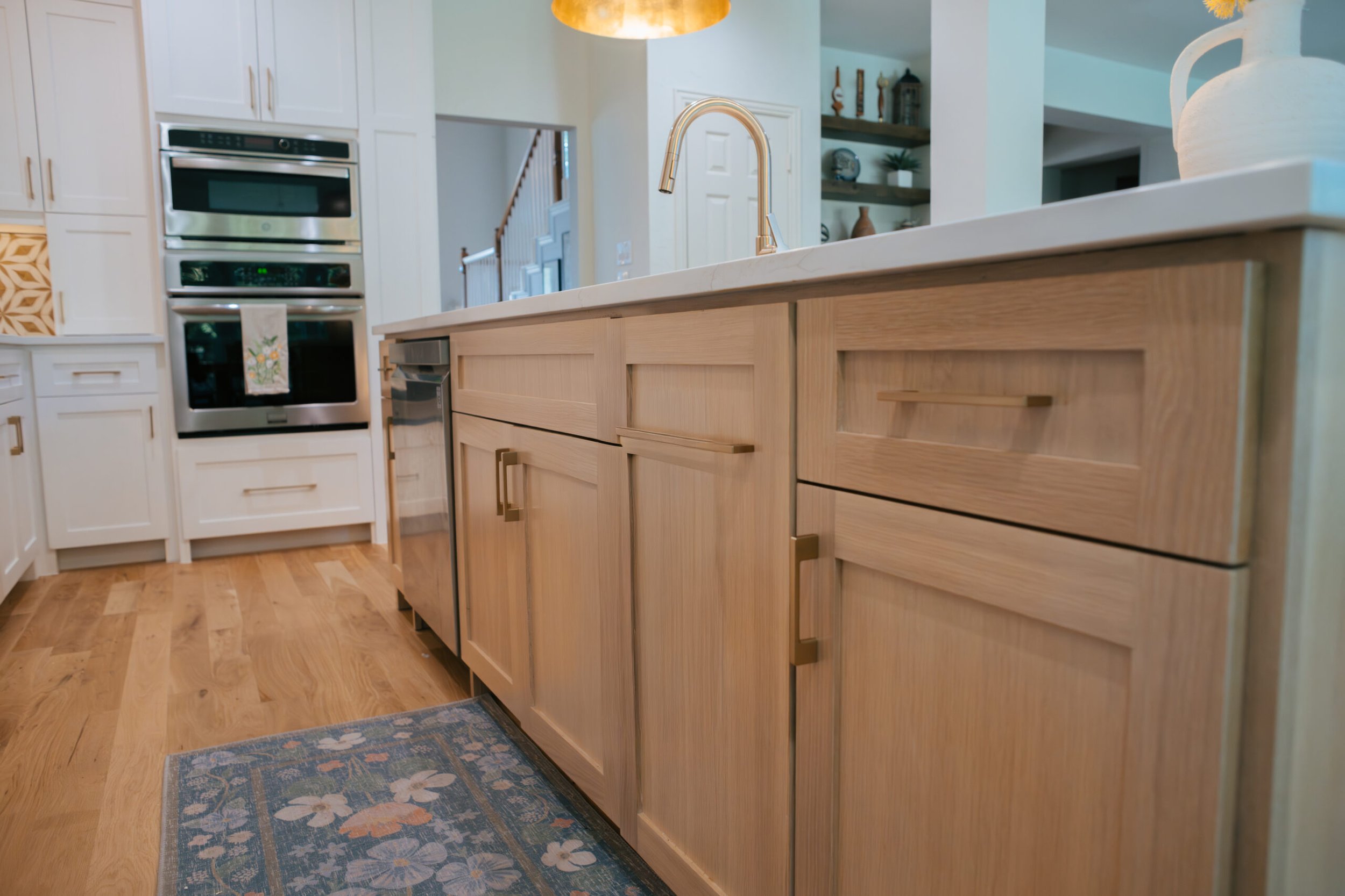 A kitchen featuring a light wooden island with cabinet drawers and doors, a white countertop, a gold-colored faucet, white cabinetry, stainless steel appliances including an oven and microwave, a decorative rug with floral patterns on wooden flooring