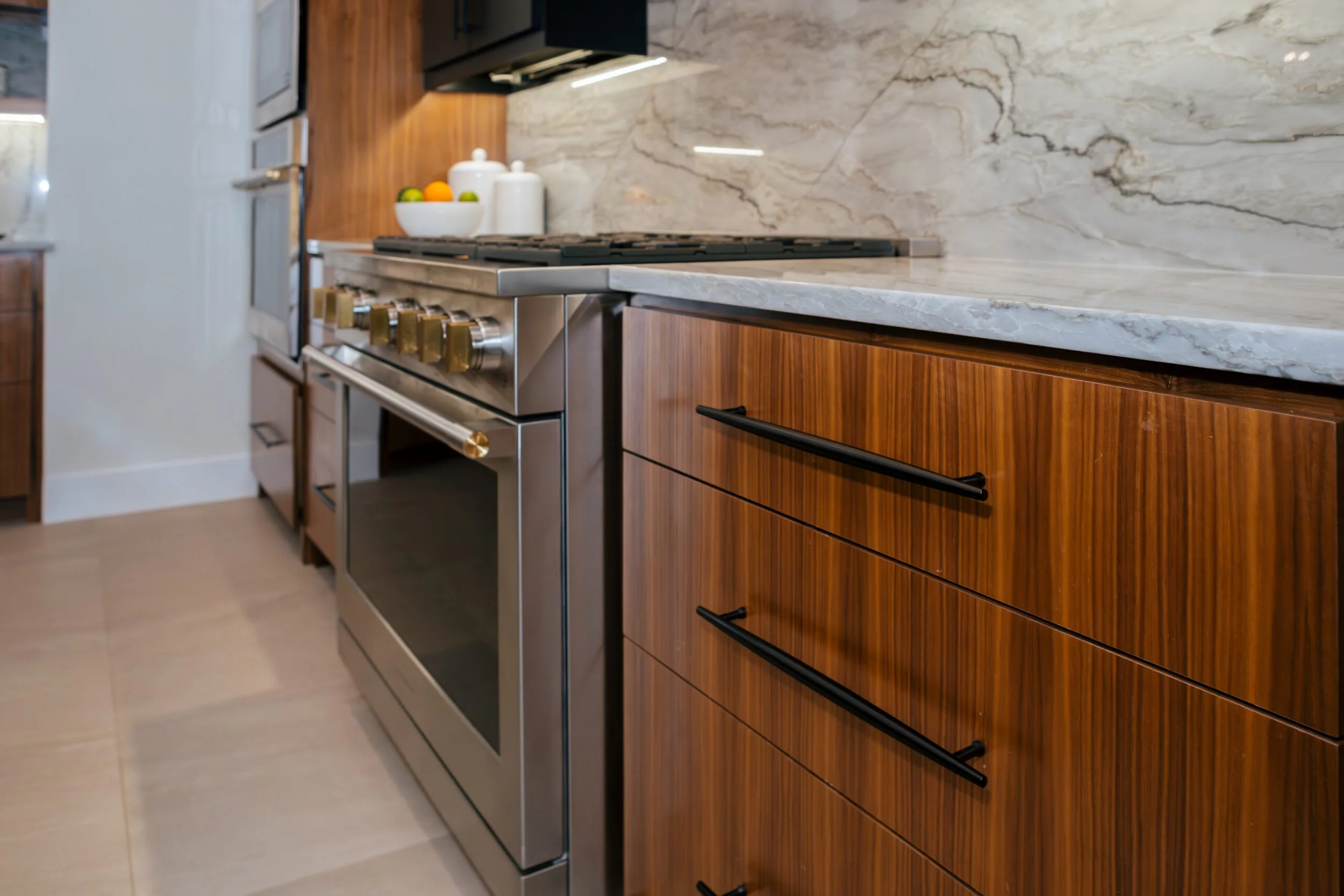 Modern kitchen with stainless steel oven and stove, dark wood cabinets, marble countertop and backsplash, and a fruit bowl with oranges and green apples.