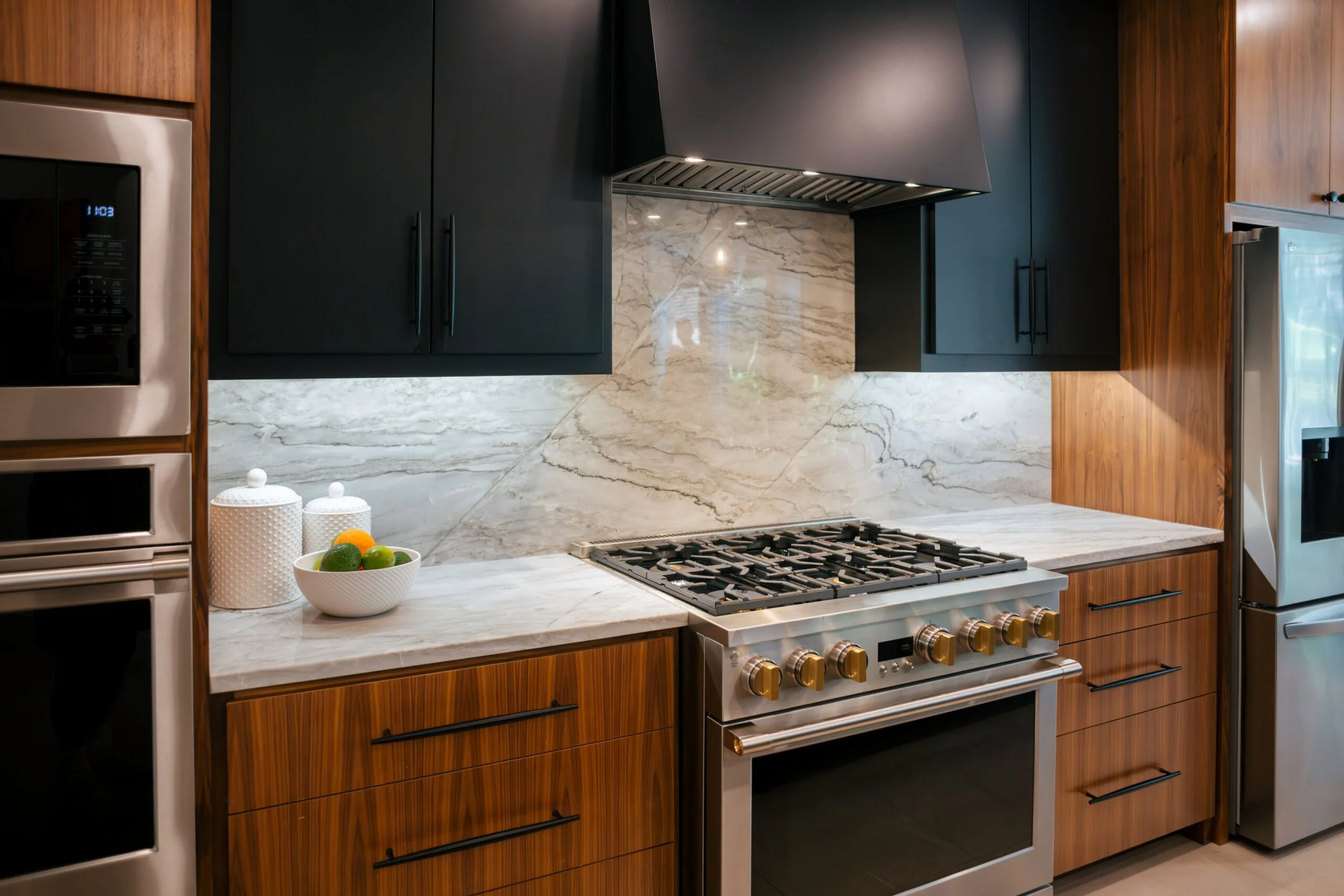 Modern kitchen with black upper cabinets, a marble backsplash, granite countertop, and stainless steel appliances, including a gas stove with gold knobs and a built-in microwave.