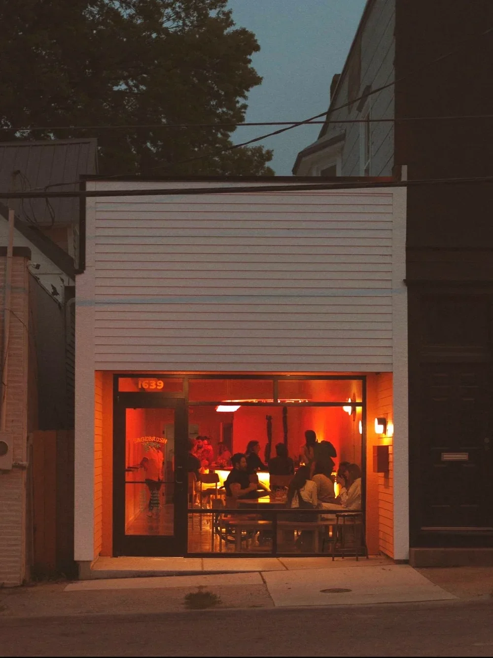 A view of a small restaurant or bar with a bright orange and red interior, seen through large glass windows at dusk. People are seated inside and the building has a modern exterior design.