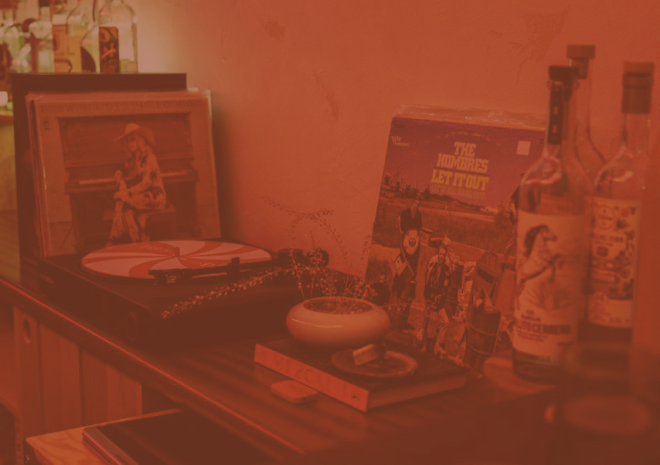 A vintage turntable with a photo of a woman playing piano leaning against the wall, a vinyl record album titled 'The Rumors of Wayout' featuring a group of skeletons, and glass bottles with colorful labels on a wooden surface.