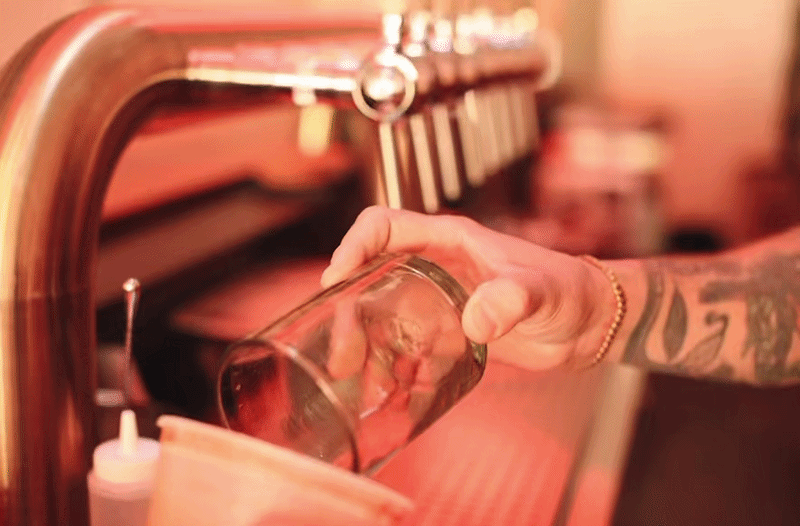 A person with tattoos and a bracelet pours a beer from a tap into a glass at a bar or brewery.
