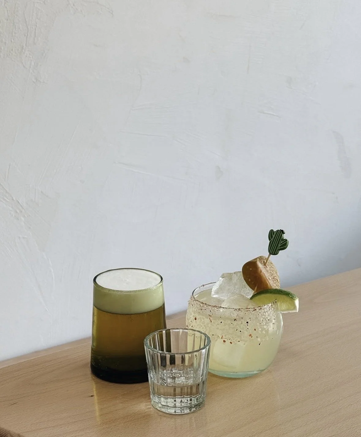 A wooden table holding three glasses: a large yellow-green drink with foam, a small clear glass of water, and a clear glass of a translucent yellow cocktail garnished with a slice of lime, a piece of ice, and a small decorative cactus. The background is a plain white wall.