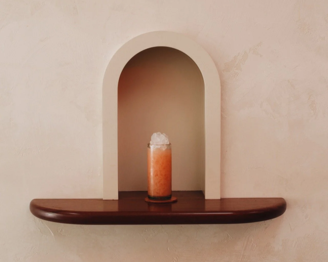 Decorative wall shelf with a centered glass candle holder containing an orange candle topped with white wax, against a textured beige wall.