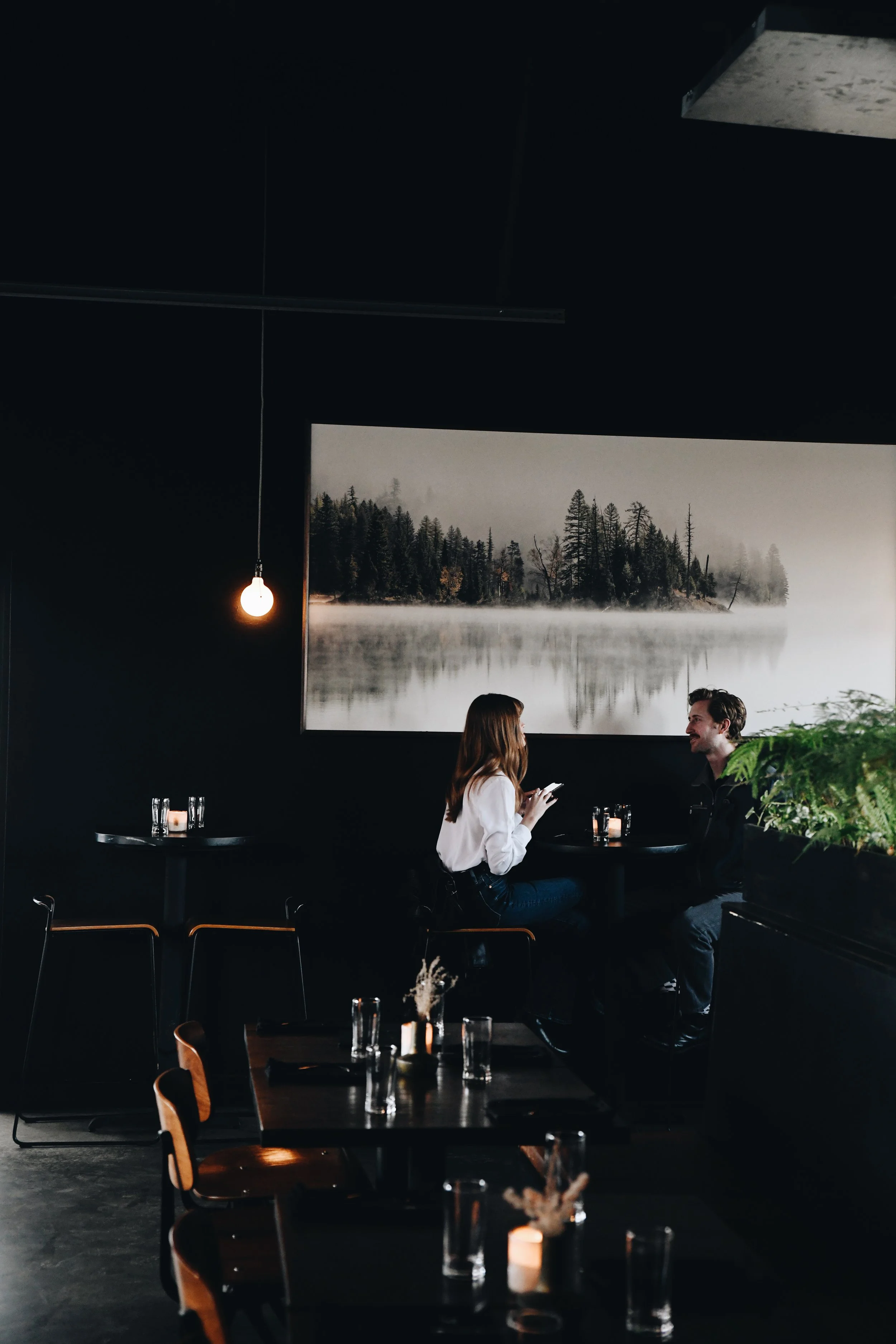 A dimly lit restaurant with dark walls, featuring a large landscape photograph of a foggy lake with trees in the background. Two people, a woman and a man, are seated at a table conversing. The woman is holding a phone, and there are glasses and candles on the tables.