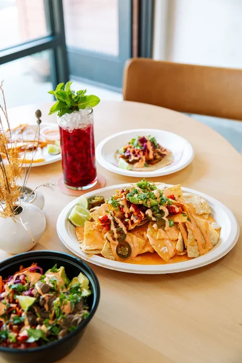 A table with Mexican food including nachos topped with cheese, jalapenos, diced tomatoes, and cilantro, a bowl of salad, and a drink with fresh mint outdoors by a window.