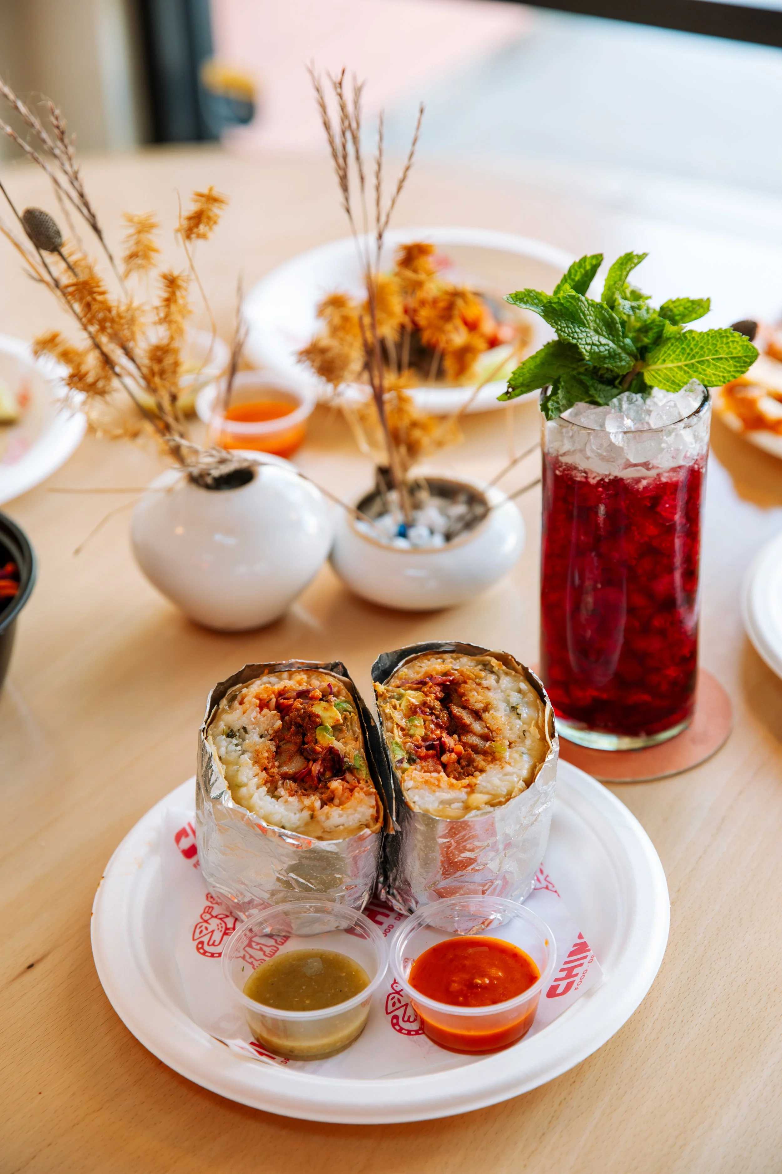 Close-up of two beef and rice burritos wrapped in foil on a plate, with two small cups of green and red dipping sauces nearby, placed on a tray at a restaurant.