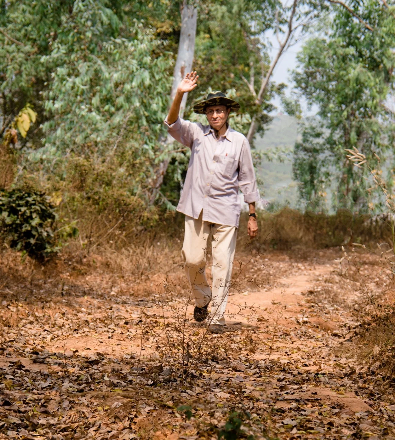 A man walking outdoors on a dirt path, wearing a hat and casual clothes, raising his hand in a wave, with green trees in the background.