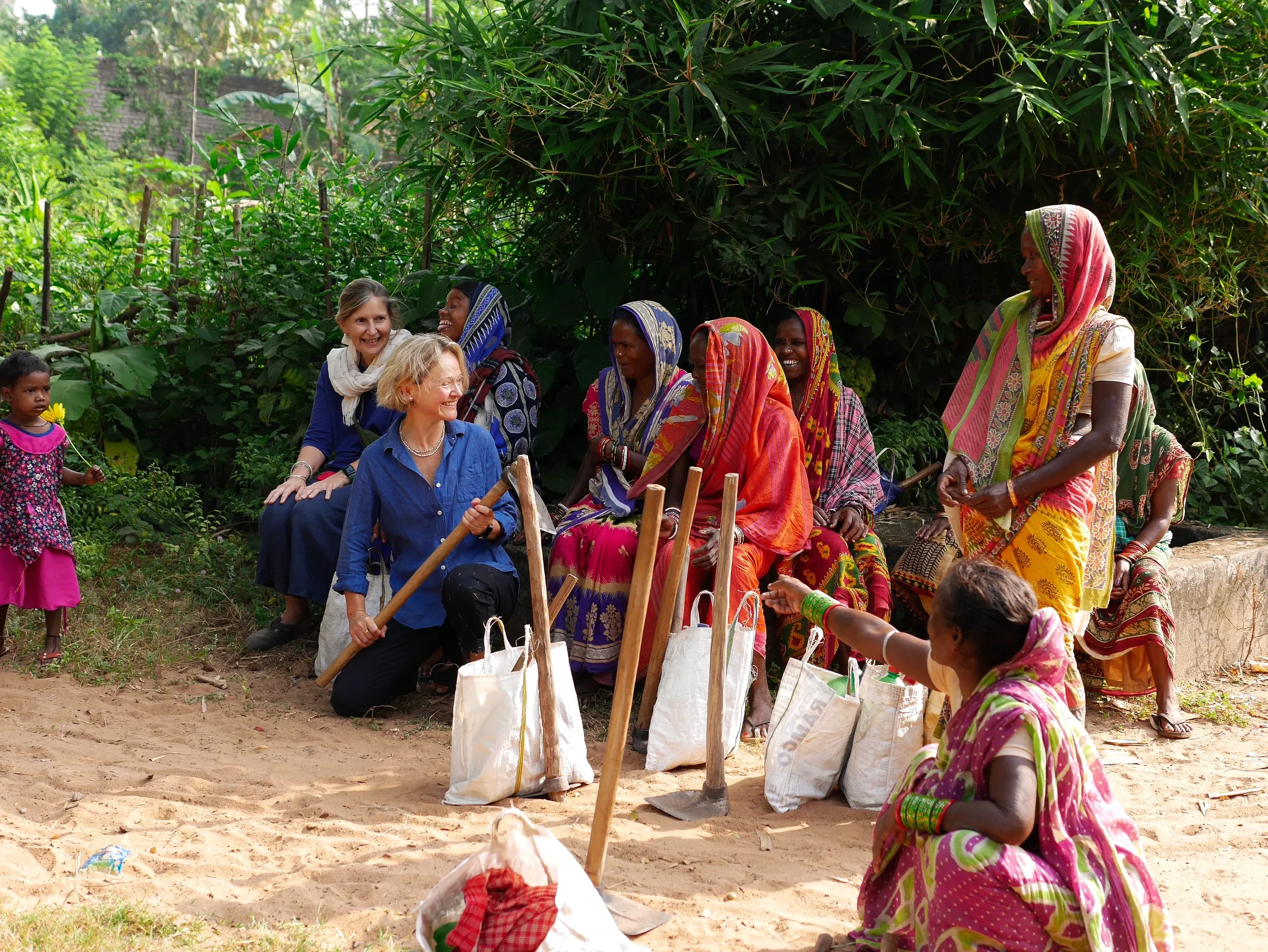 Women and children sitting and standing outdoors in a lush green area with trees, engaging in conversation and smiling.
