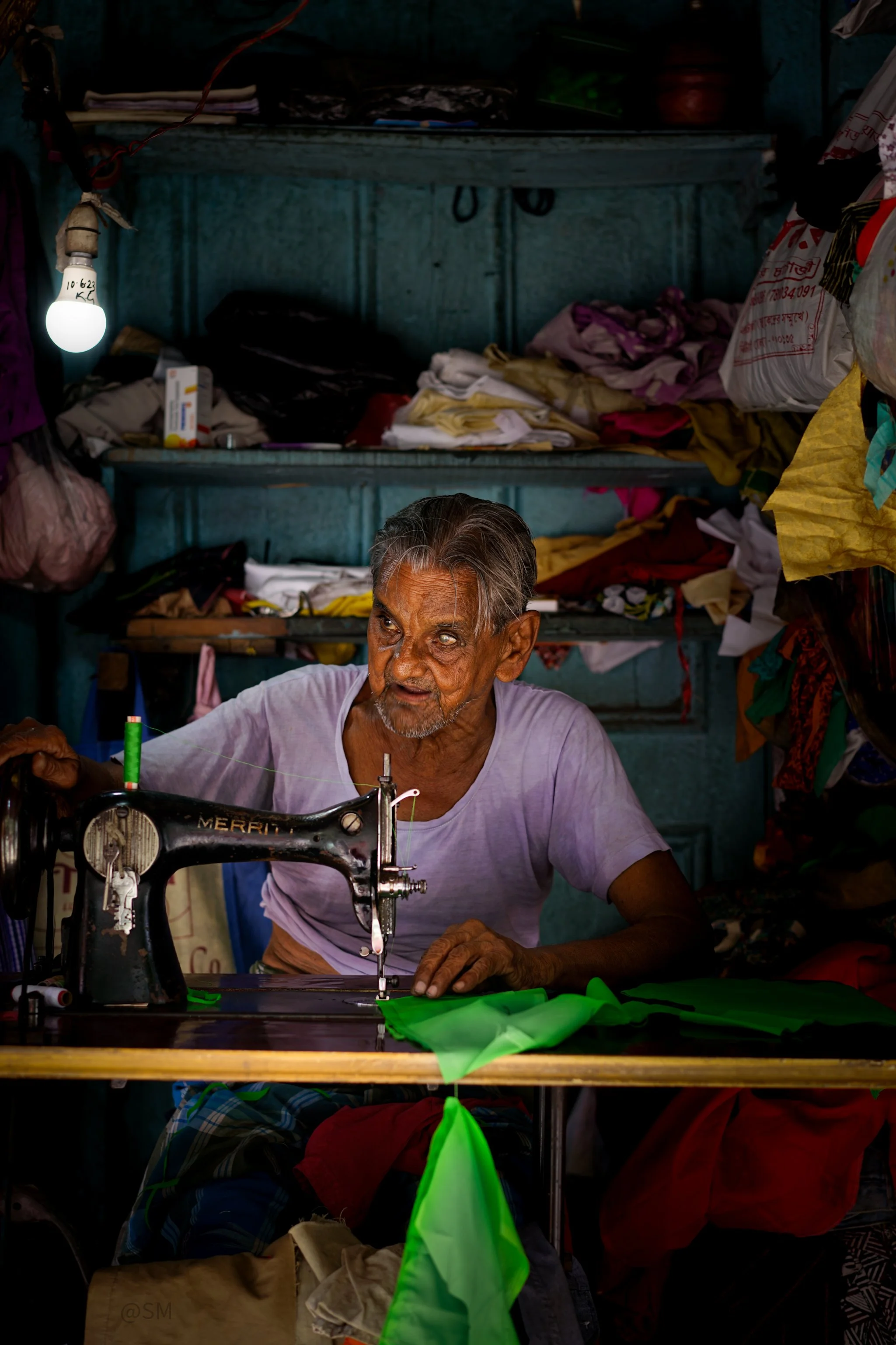 An elderly man with gray hair and a beard is working on a sewing machine amidst colorful fabrics and cluttered background with shelves and bags.