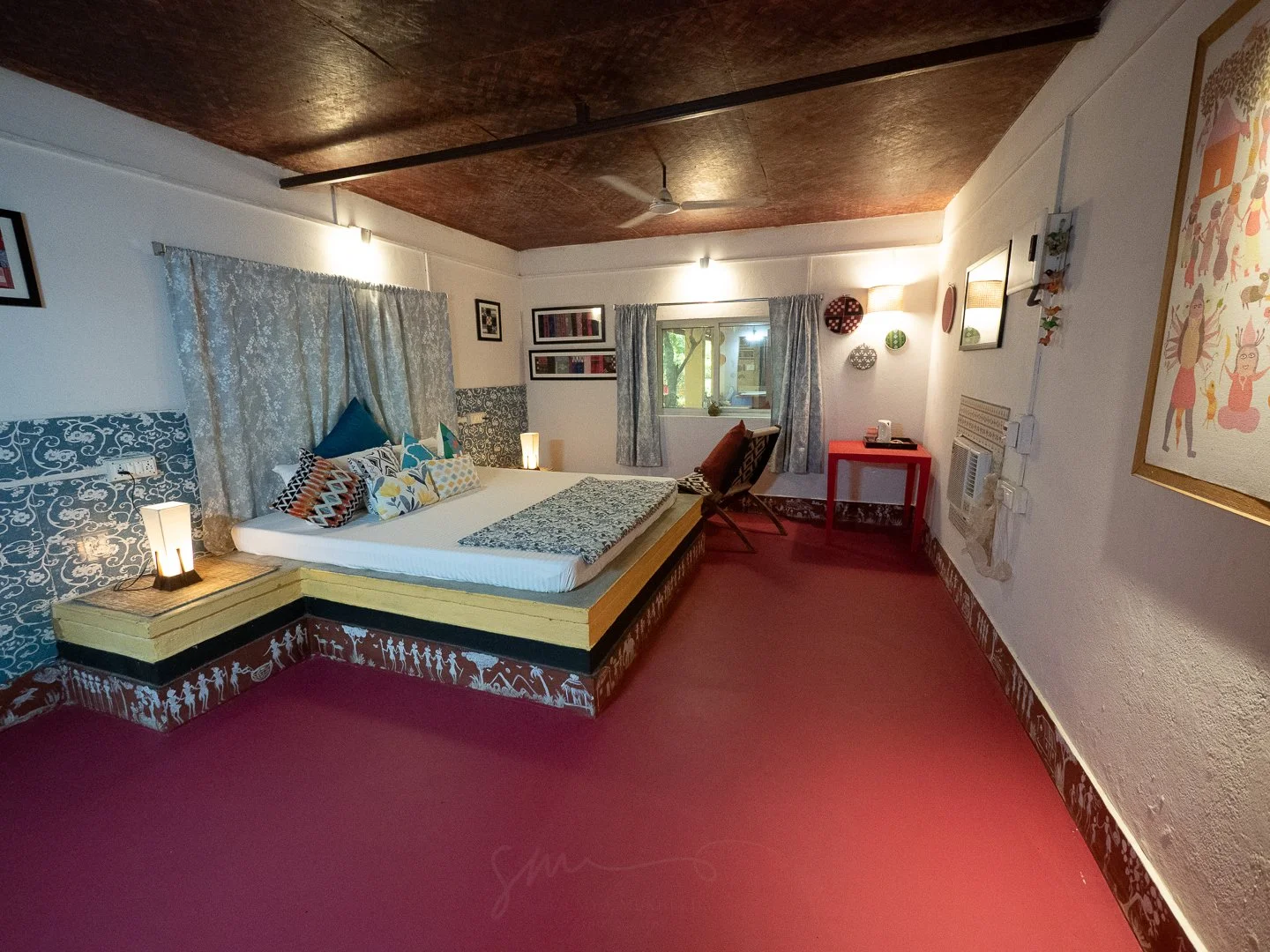 Comfortable bedroom with a bed featuring patterned pillows, decorated with lace curtains and artwork on the walls, and a small window revealing greenery outside.