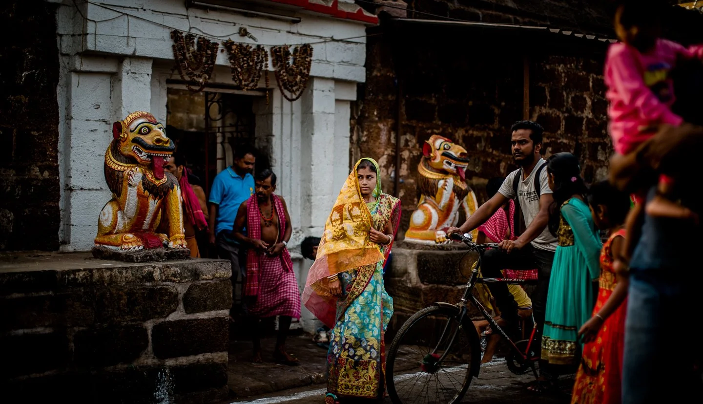 A woman dressed in traditional Indian attire standing near a man on a bicycle surrounded by other people, with colorful lion statues in the background.