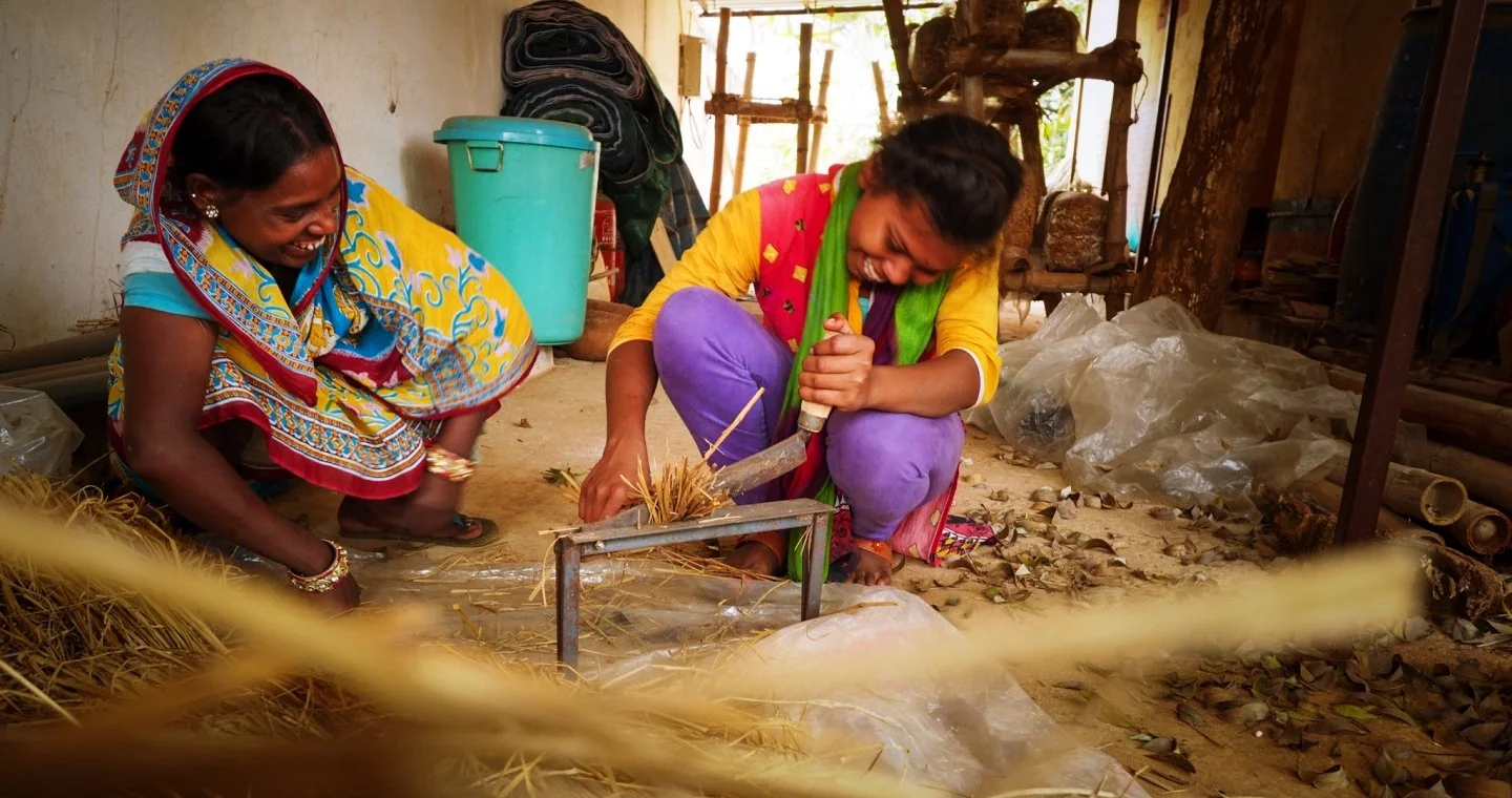 Two women in colorful traditional clothing sitting on the ground, working with straw or grass in a rustic room. One woman is using a hand tool, possibly for weaving or crafting.