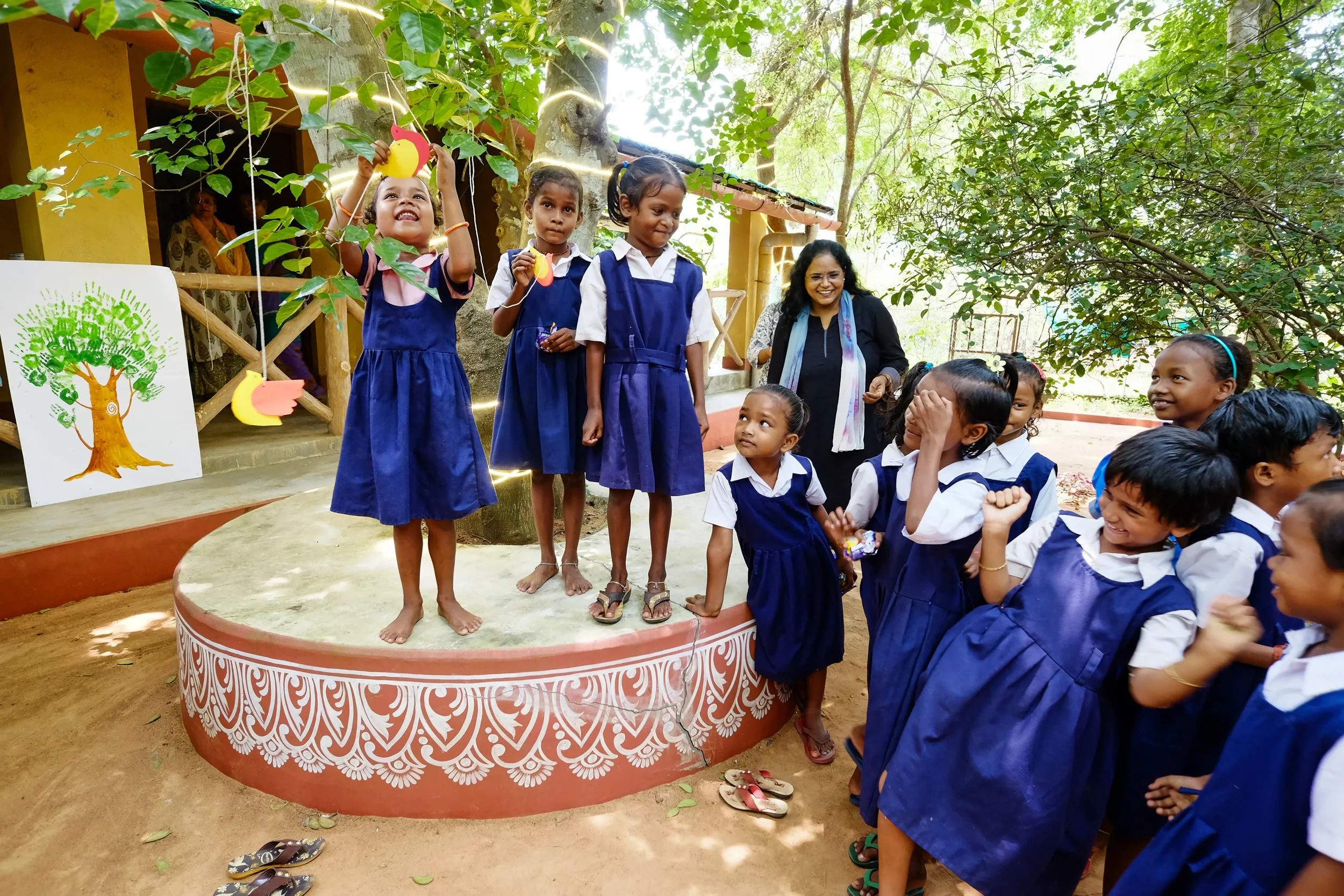 Children wearing blue school uniforms are gathered around a small stage outdoors, with some children on the stage and others in the audience, under the shade of trees. The children are smiling, and one girl is holding a colorful paper craft. There is a woman in black with a scarf smiling nearby, and a large drawing of a tree is visible behind the children on stage.