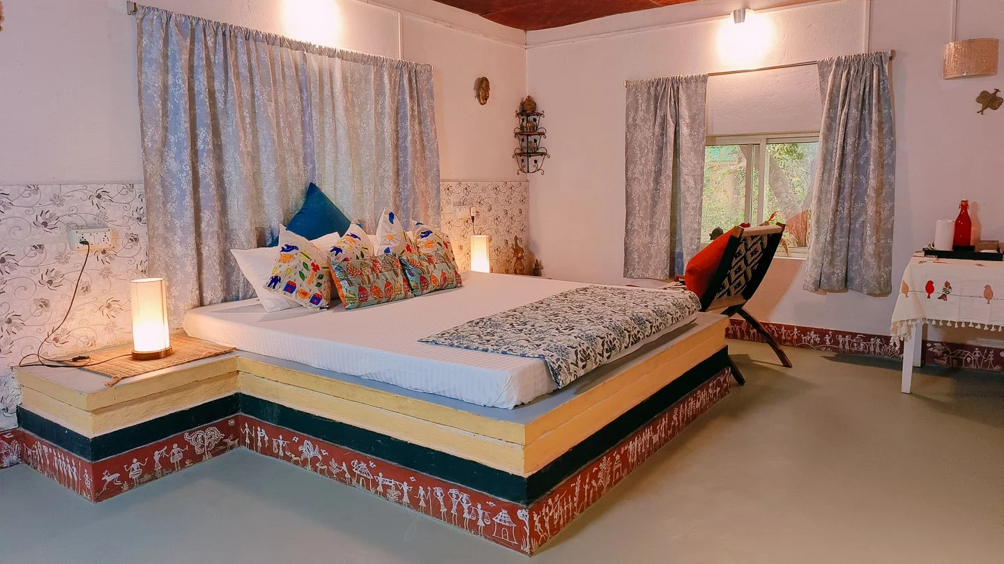 A bedroom with a large bed featuring colorful pillows and a patterned throw, two lamps on either side, a chair with an orange pillow, a window with curtains, and a small table with decorative items.