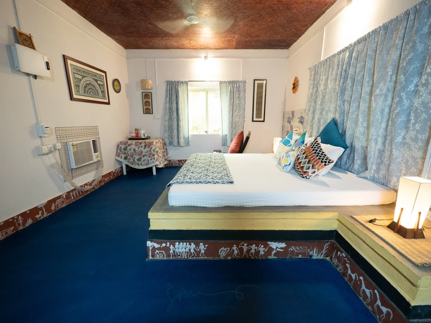 A cozy bedroom with a platform bed decorated with patterned pillows and light blue curtains. The bed is elevated with animal and nature-inspired designs on its base. To the left, a small table with a floral tablecloth holds a microwave and other small appliances. The room has a window with natural light, wall art, and a string of decorative items. The ceiling is wooden, and the floor is covered in dark blue carpet.