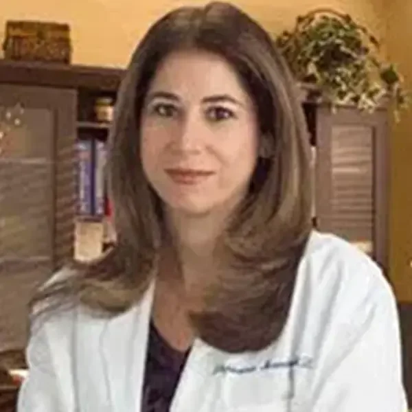 A woman with shoulder-length brown hair wearing a white lab coat, sitting in a room with bookshelves and a plant in the background.