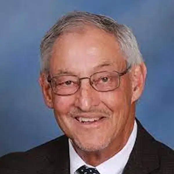 Headshot of an older man with gray hair and glasses, wearing a suit and tie, smiling against a blue background.