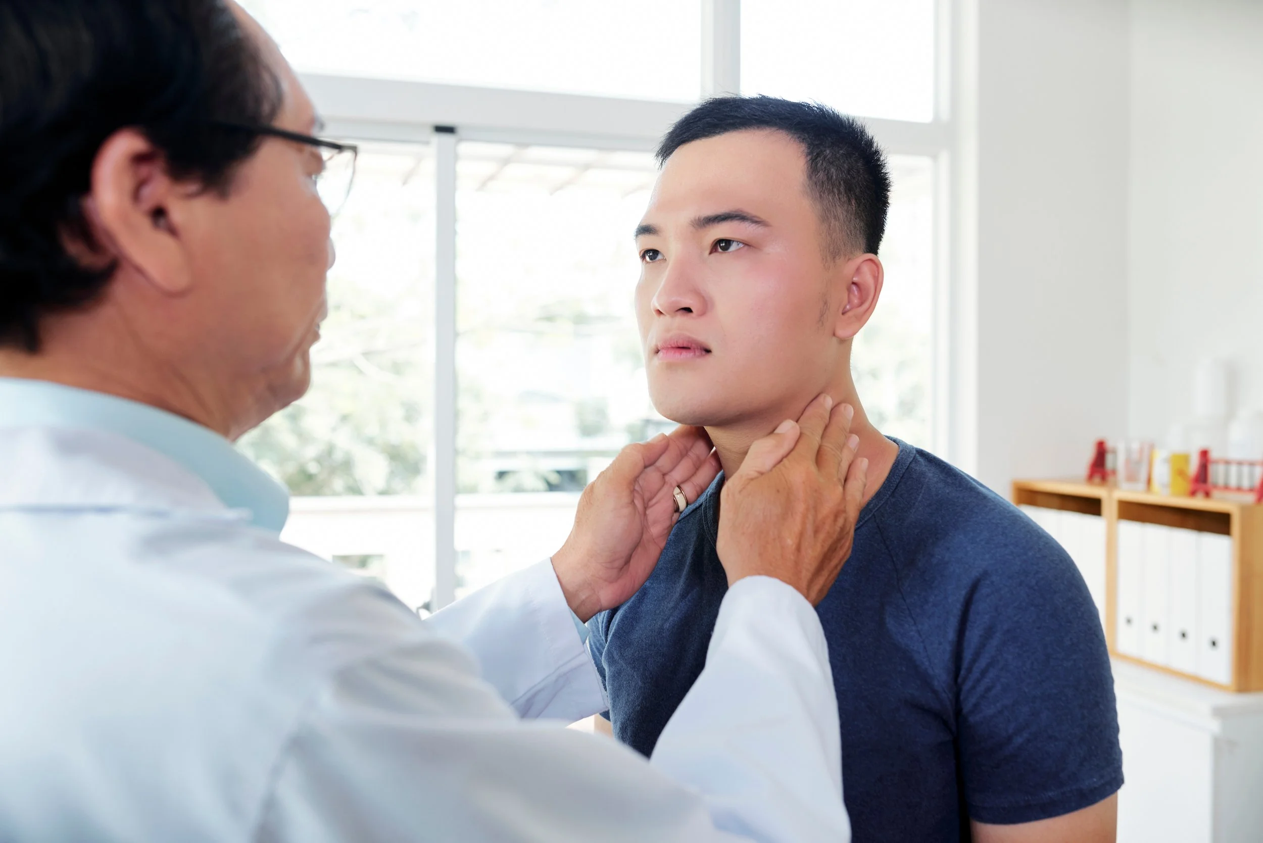 A healthcare professional examines a young man by gently holding his chin and neck, both individuals are focused on the examination in a bright room with large windows.