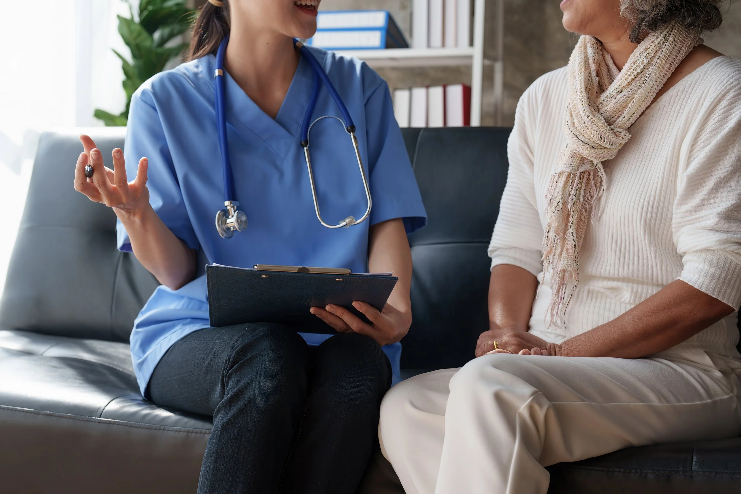 Medical professional in blue scrubs talking with an older woman in a white sweater, sitting on a gray couch in a bright room with shelves and green plant in background.
