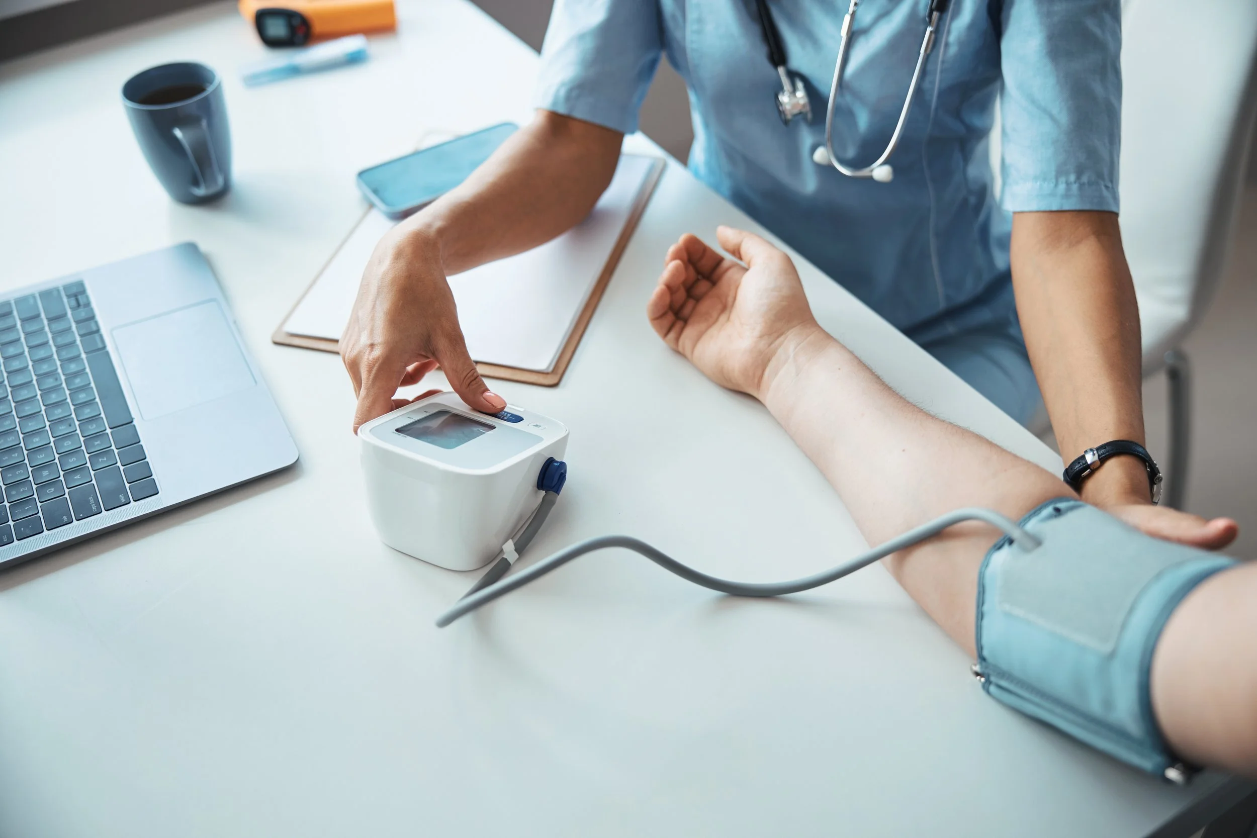 Medical professional wearing a stethoscope takes a person's blood pressure using a digital blood pressure monitor at a white table with a laptop, coffee mug, and notepad.