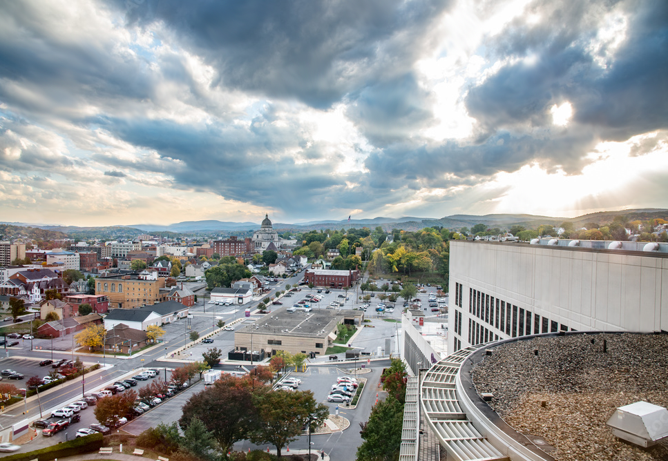 A cityscape view with a mix of modern and historic buildings, parking lots, and trees, under a dramatic cloudy sky with rays of sunlight.