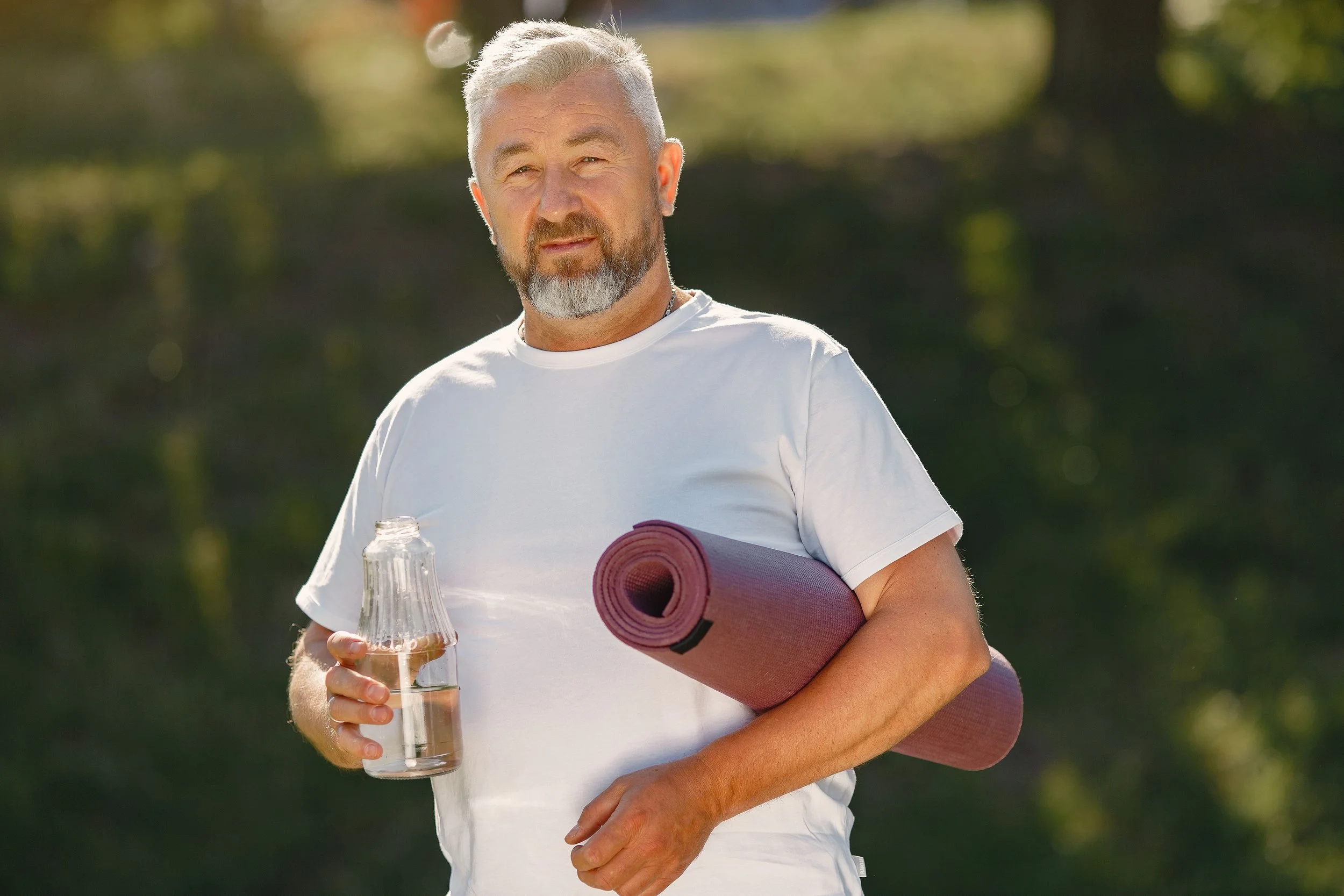 Older man with gray hair and beard holding a glass water bottle and a rolled yoga mat outdoors.
