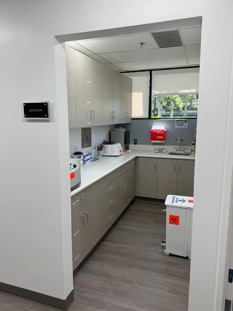 Image of a small laboratory room with beige cabinets, countertop, lab equipment, and a window showing trees outside.