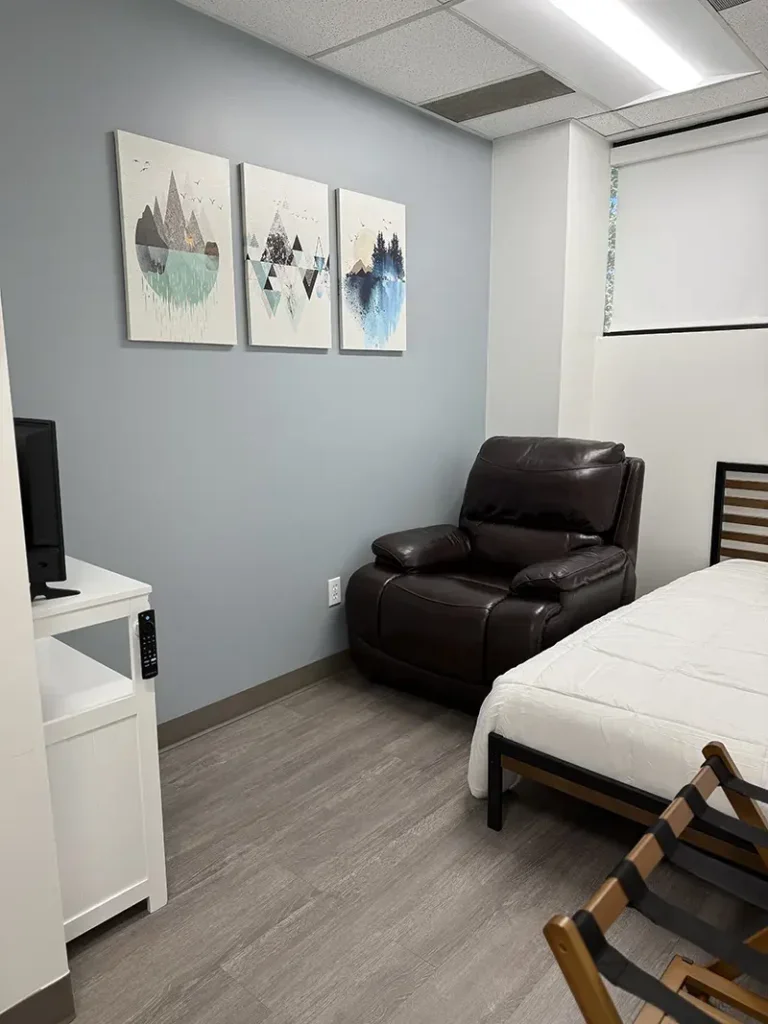 A corner of a bedroom with a white bed, a black reclining chair, a white TV stand with a small TV, and three modern landscape paintings hanging on a light blue wall.