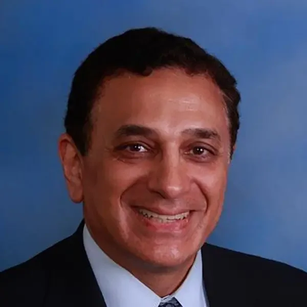 A smiling man with short dark hair, wearing a dark suit and tie, against a blue background.
