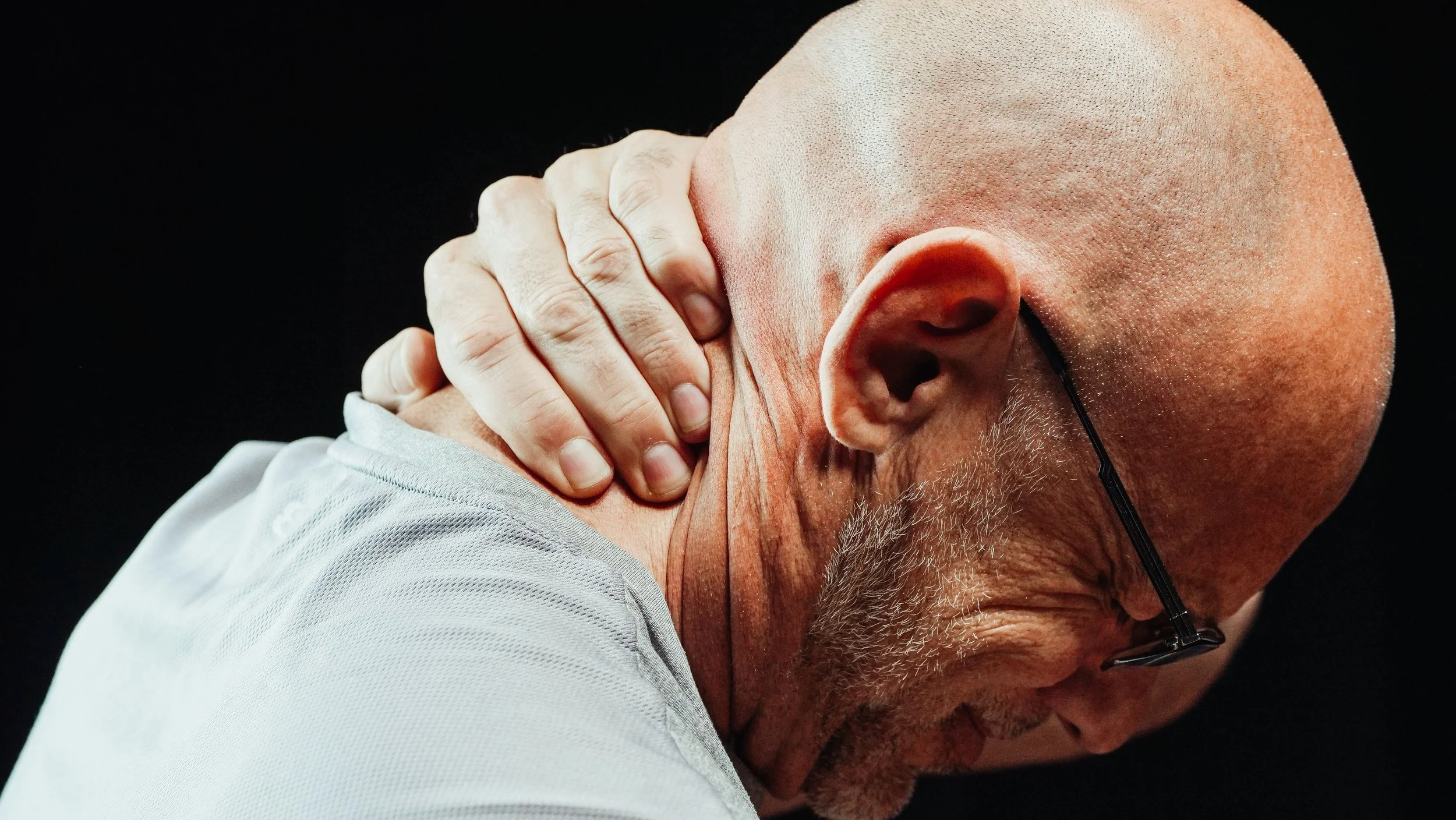 A bald man with glasses and a beard, touching his neck with one hand, against a black background.
