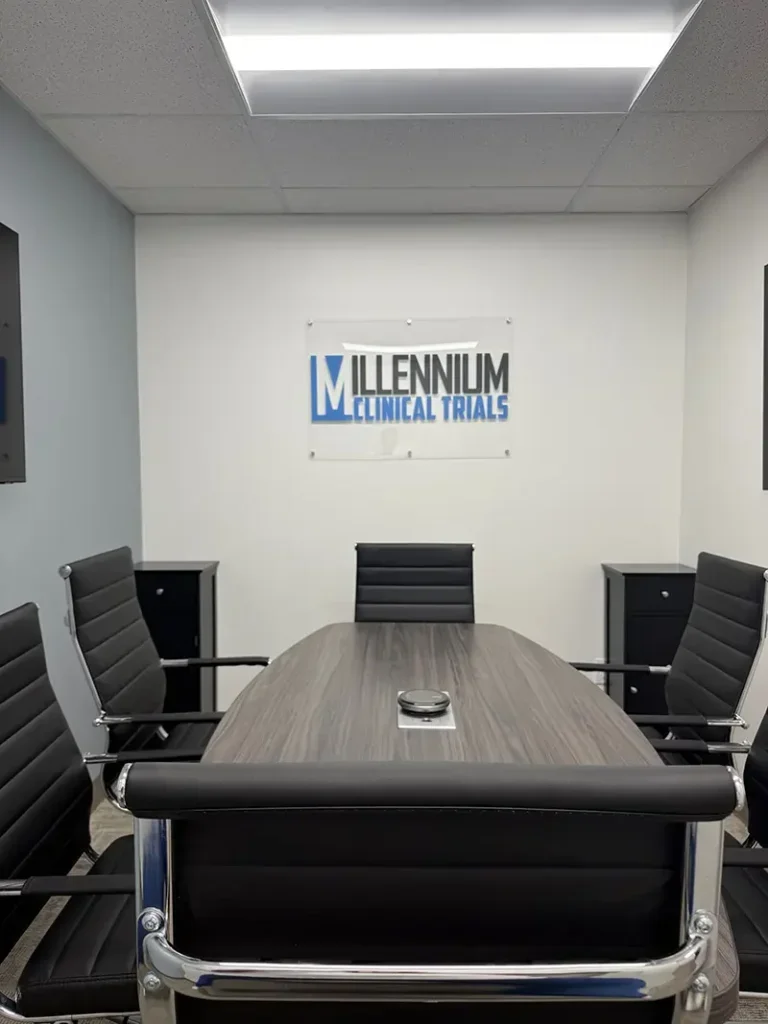 A conference room with a wooden table surrounded by five black office chairs, a sign on the wall that reads 'Millennium Clinical Trials', and a light-colored wall and ceiling with fluorescent lighting.