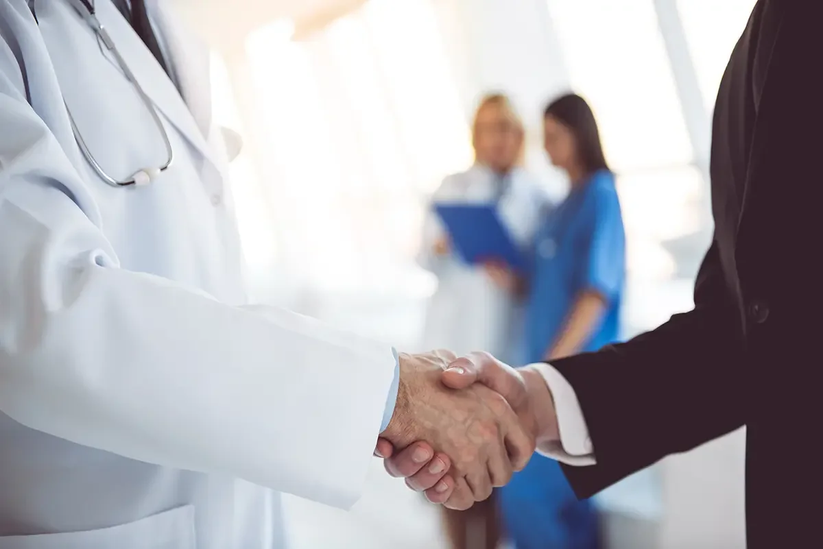 Doctor and businessman shaking hands in a medical setting, with medical professionals in the background.