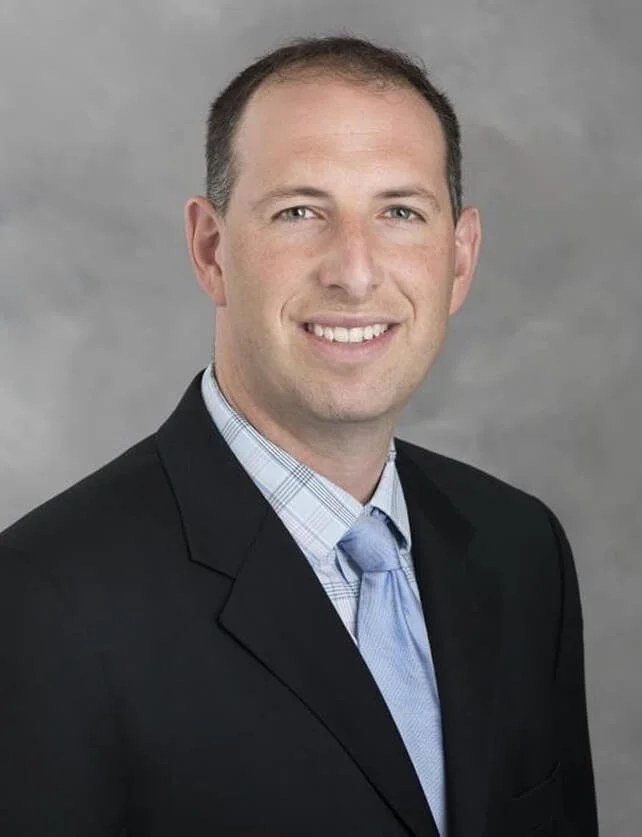 A professional headshot of a man with short brown hair, wearing a dark suit jacket, a light blue shirt, and a matching light blue tie, smiling against a neutral gray background.