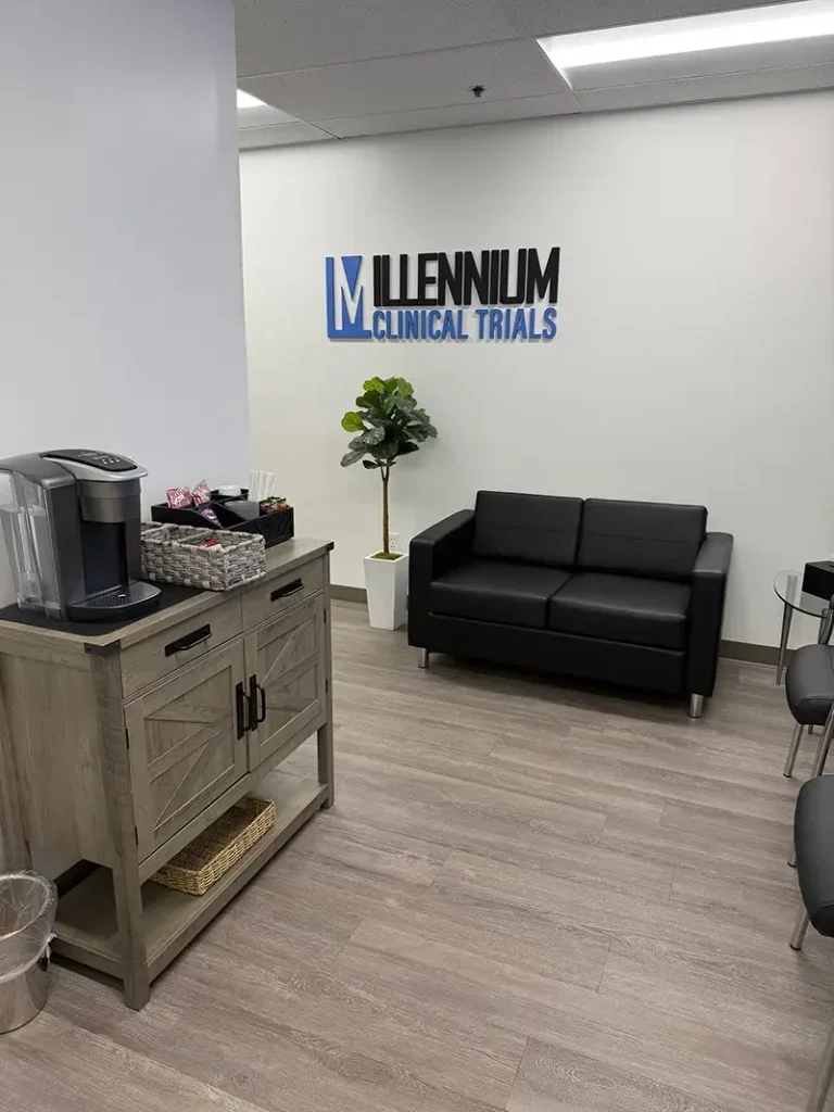 Interior of a waiting room at Millennium Clinical Trials with a coffee station, a black sofa, and a potted plant on the wall.