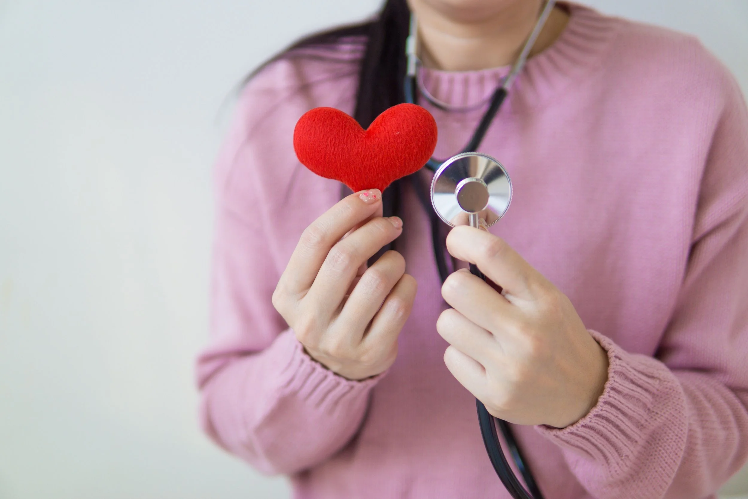 A person wearing a pink sweater holds a red plush heart-shaped object in one hand and a stethoscope near their chest with the other hand, suggesting a medical or health-related theme.