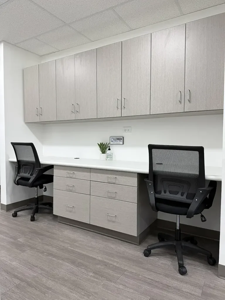 Empty office workspace with two black office chairs, a white countertop, gray cabinets above and below, a potted plant, and a wall outlet with a small decor sign.