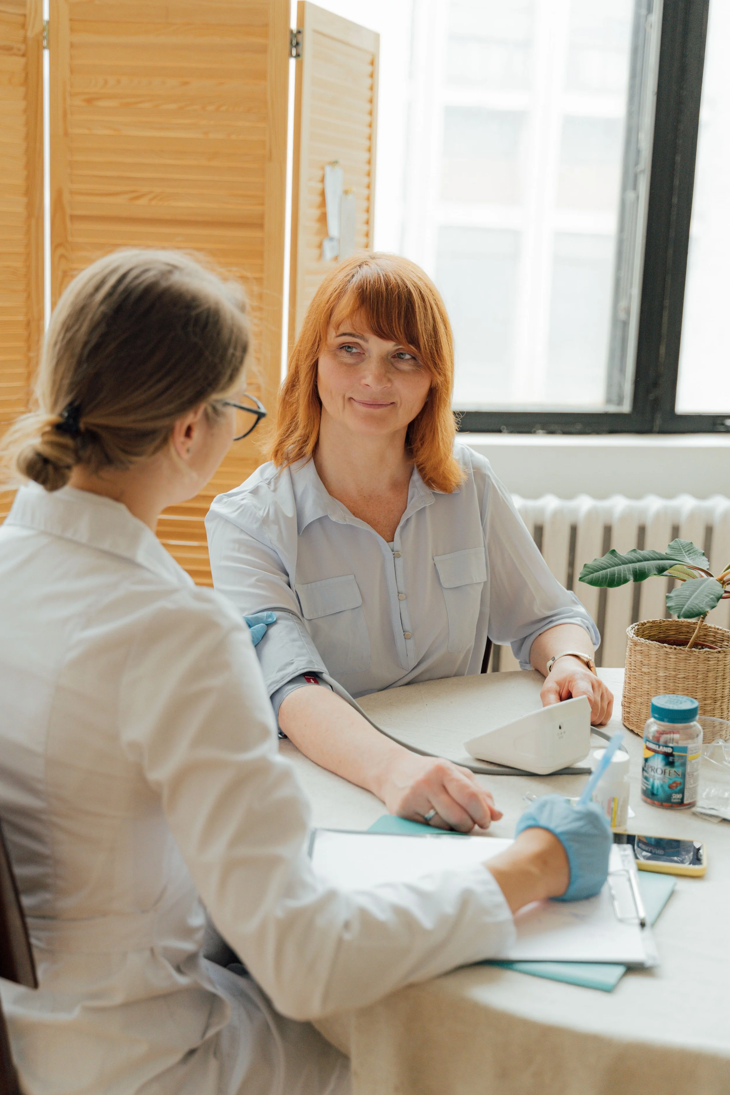 Two women in medical attire having a discussion in a well-lit room with large windows, a wooden room divider, and medical supplies on the table.