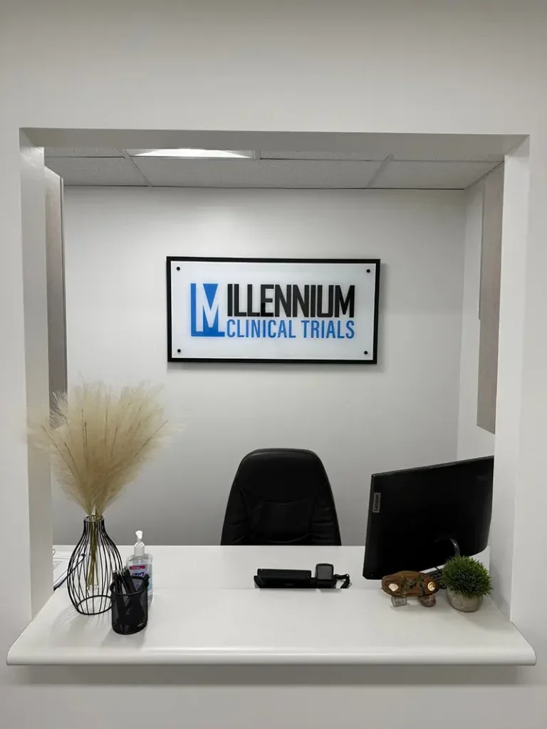 Reception desk with a black chair, computer monitor, hand sanitizer, a vase with dried pampas grass, a small plant, and personal items, with a sign that reads 'Millennium Clinical Trials' on the wall.
