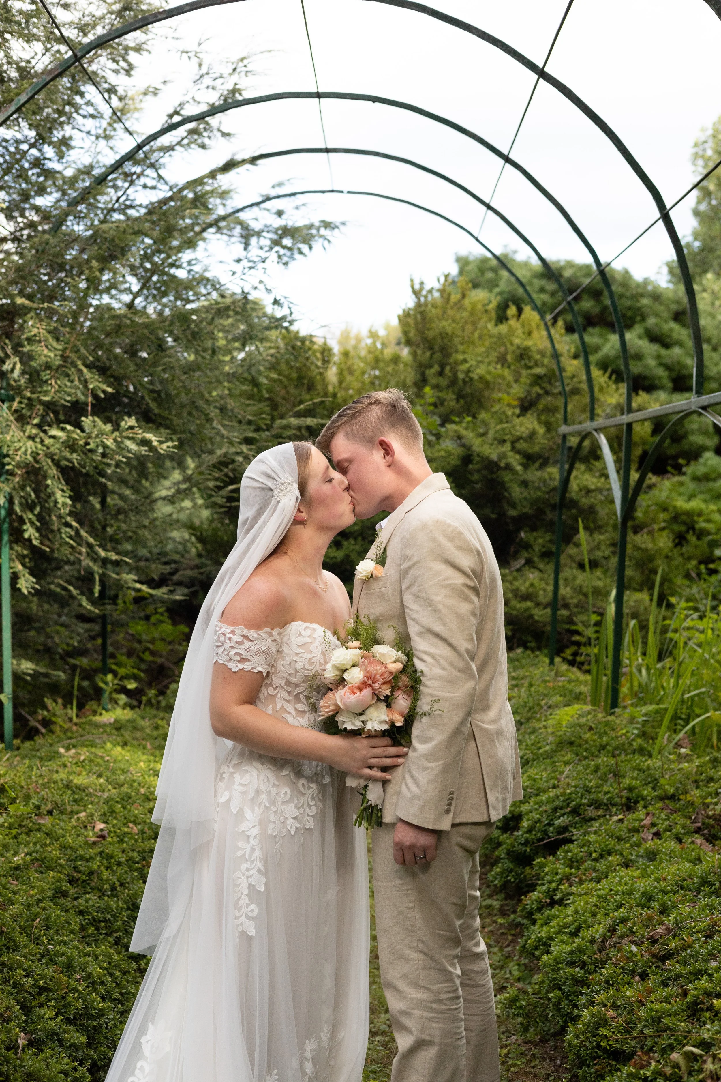 A bride and groom share a kiss under a garden archway surrounded by lush greenery during their wedding.