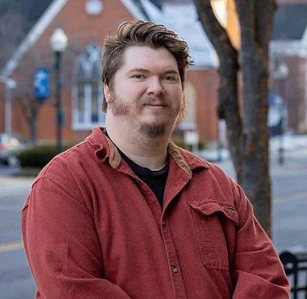 A young man with light skin, light brown hair, and beard wearing a red button-up shirt outdoors in a suburban area.