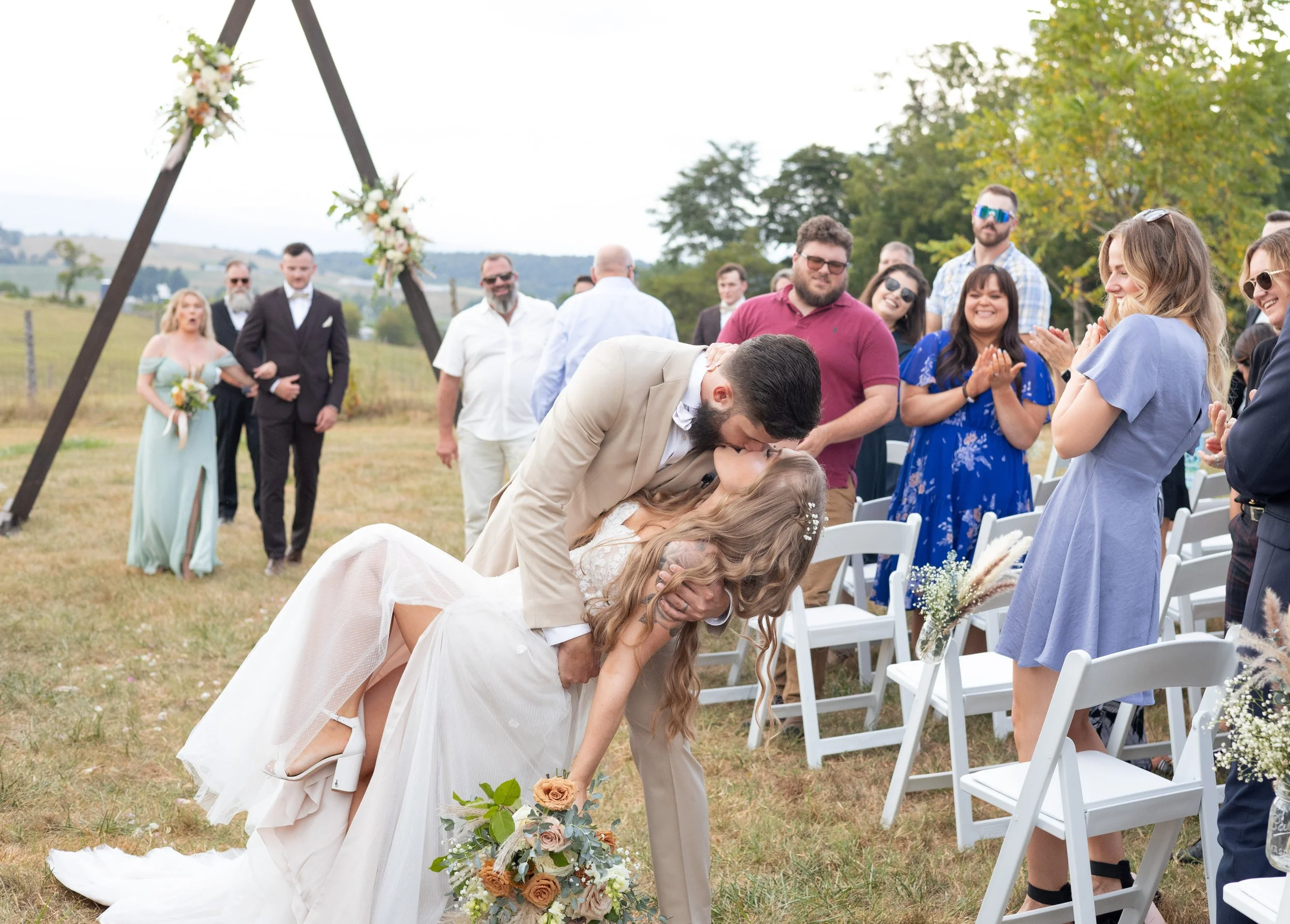 A wedding ceremony takes place outdoors, with a bride and groom leaning in for a kiss. The bride holds a bouquet of flowers, and the groom is dressed in a light-colored suit. Guests are standing and clapping, smiling, and celebrating, with a decorated arch in the background on a grassy field surrounded by trees.