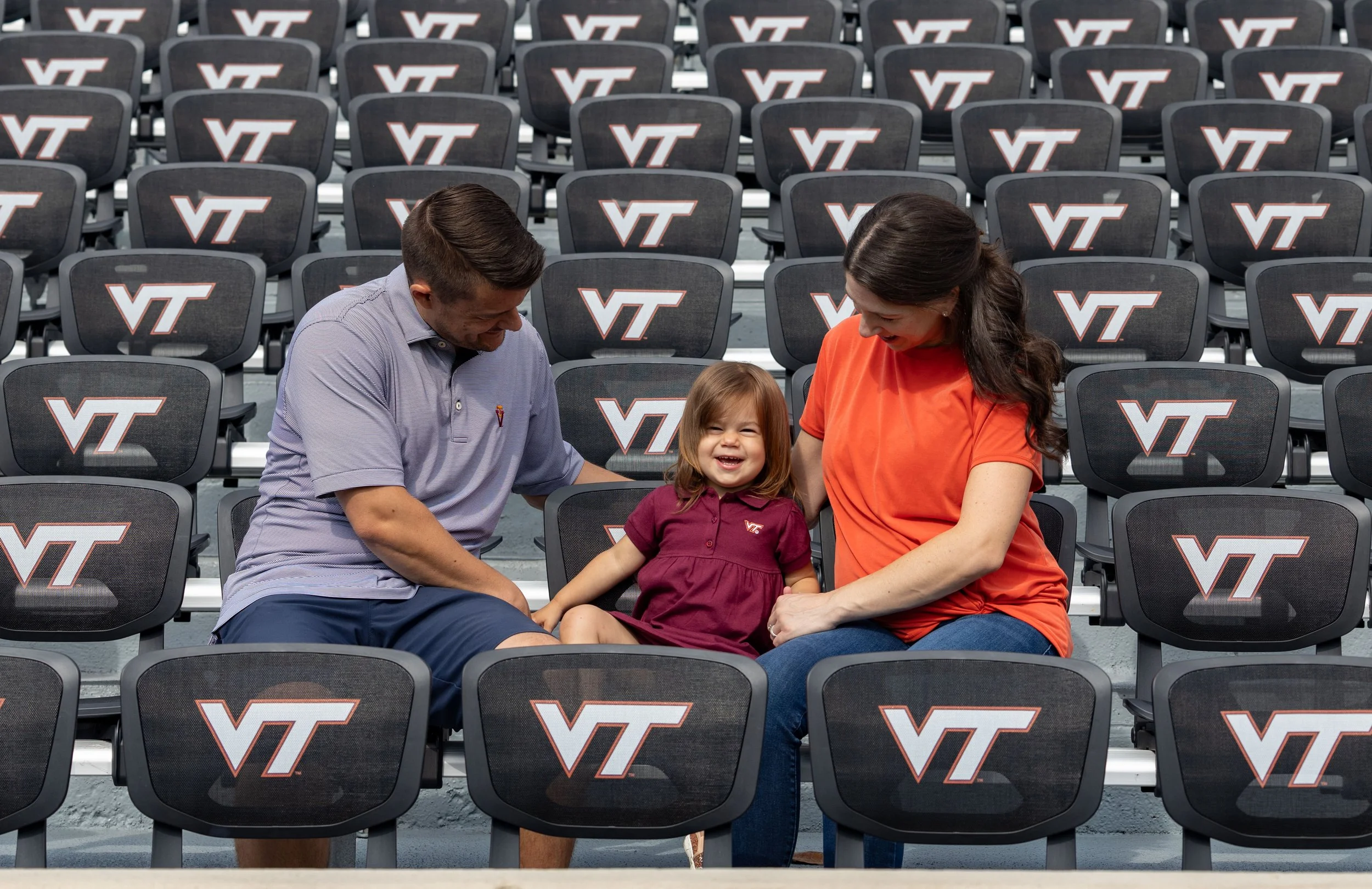 A family of three, including a young girl, seated among Virginia Tech VT branded stadium chairs. The girl is smiling at the camera while her parents look at her affectionately.