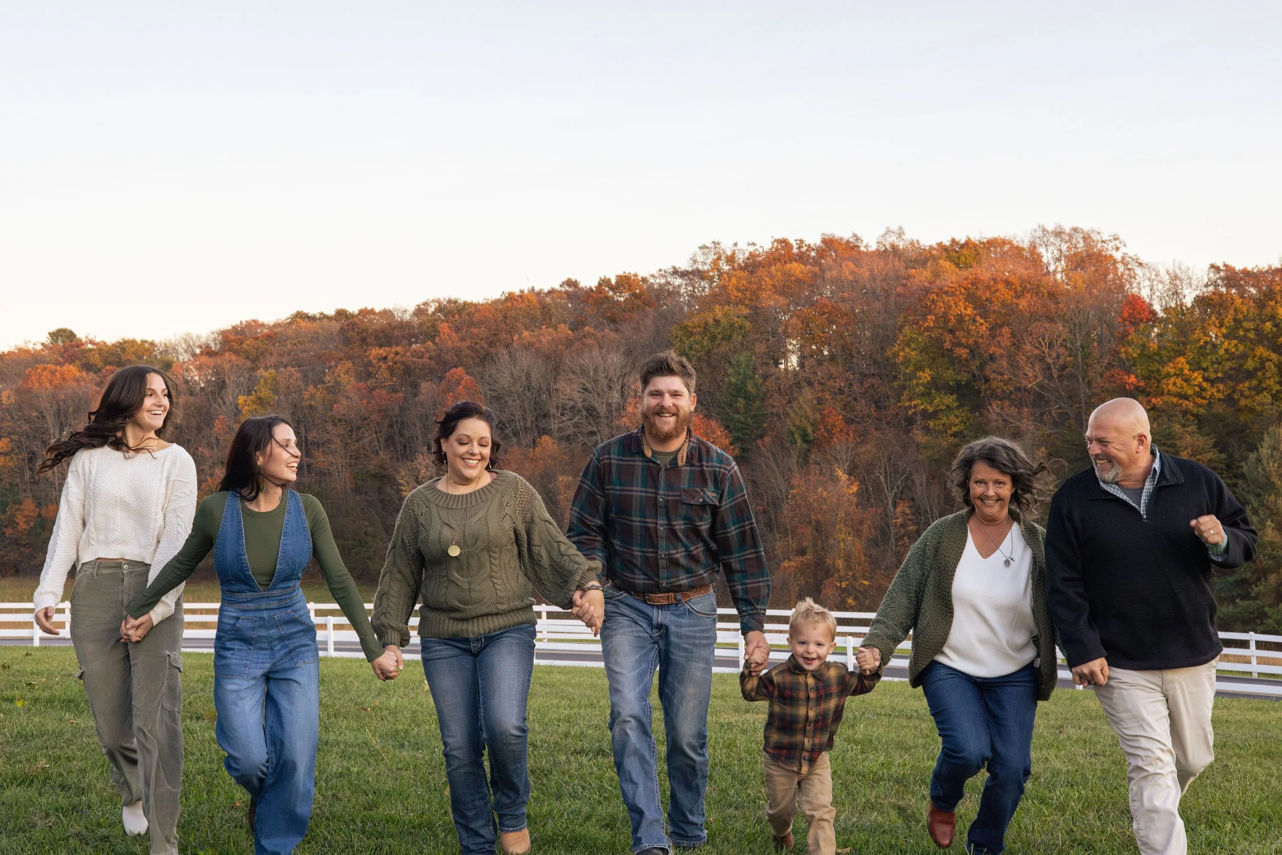 A diverse group of people, including children and adults, holding hands and walking outdoors on a grassy field with autumn trees in the background.