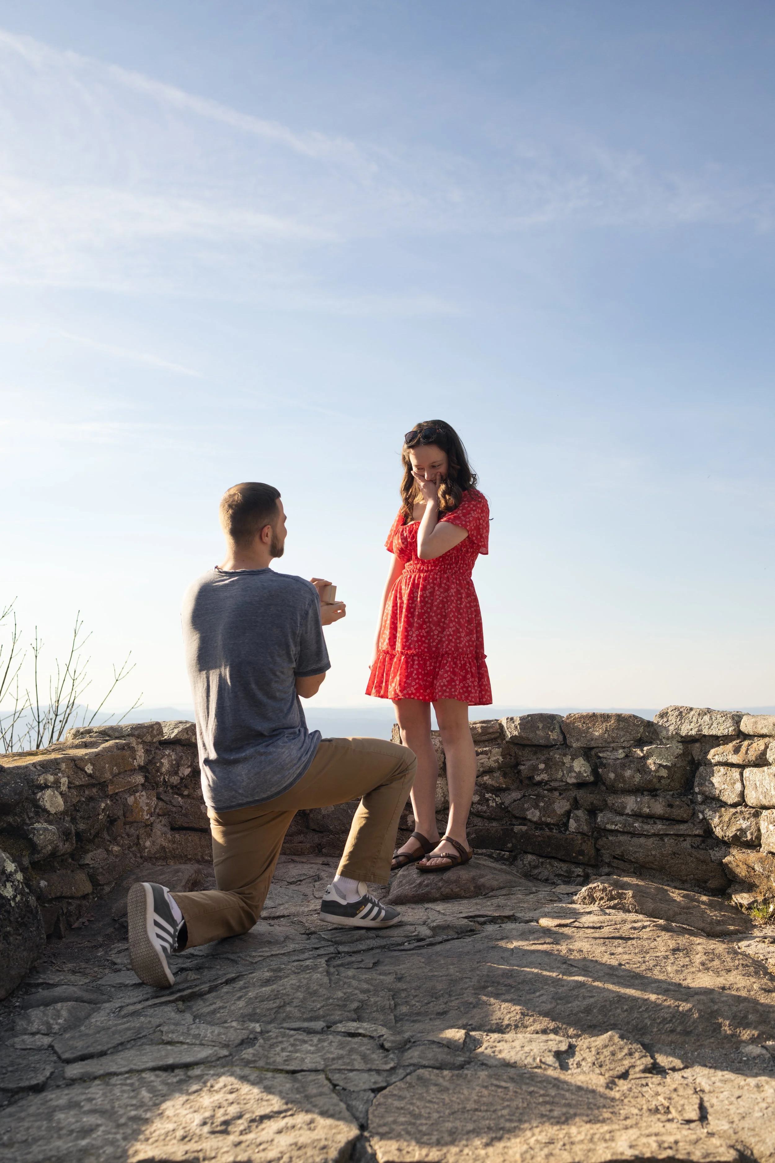 A man kneeling on a stone path proposing marriage to a woman standing in front of him, both smiling, outdoors with a clear sky.