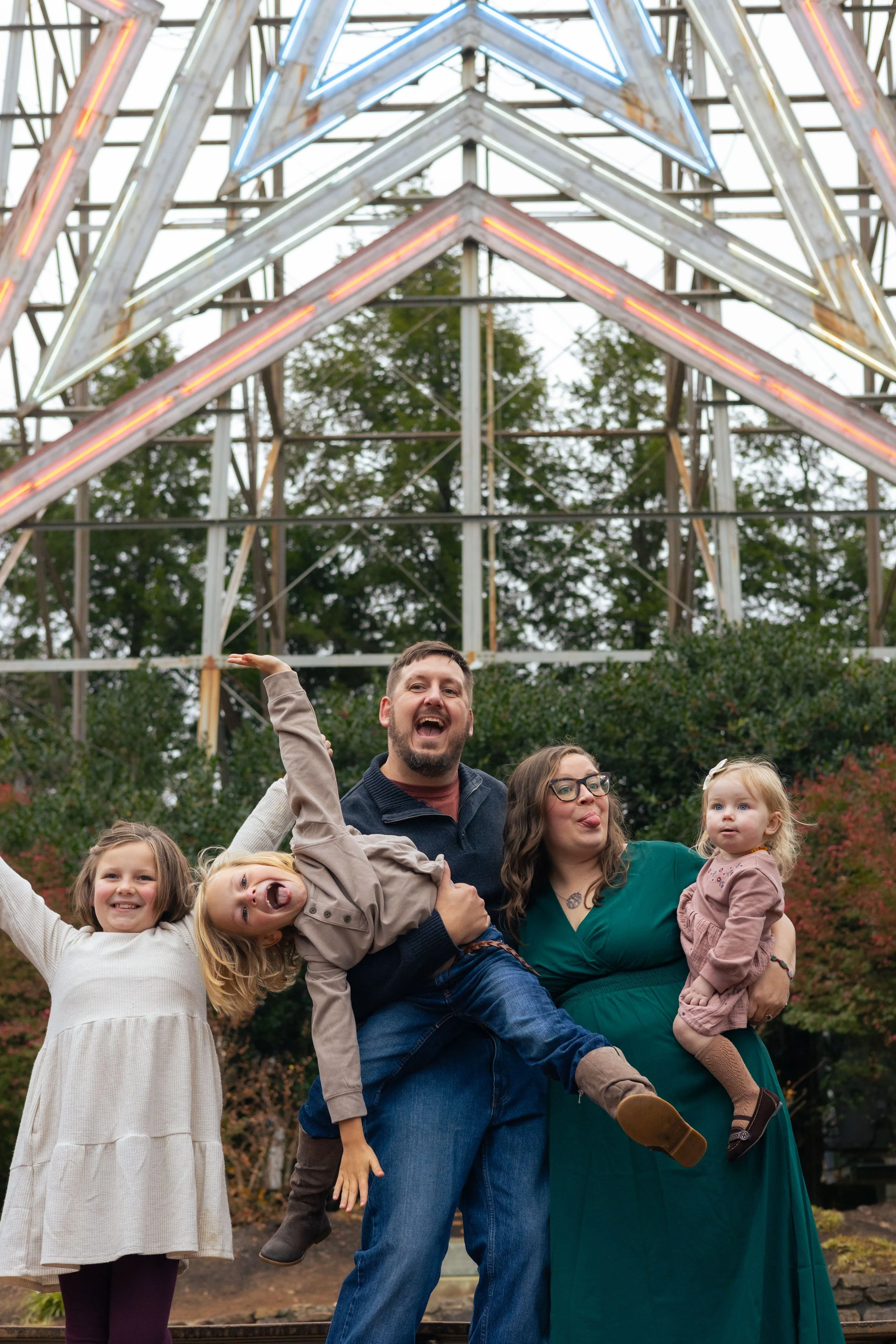 A group of smiling children and adults posing outdoors in front of a metal amusement park ride with illuminated star shapes, trees in the background, people dressed warmly, during daytime.