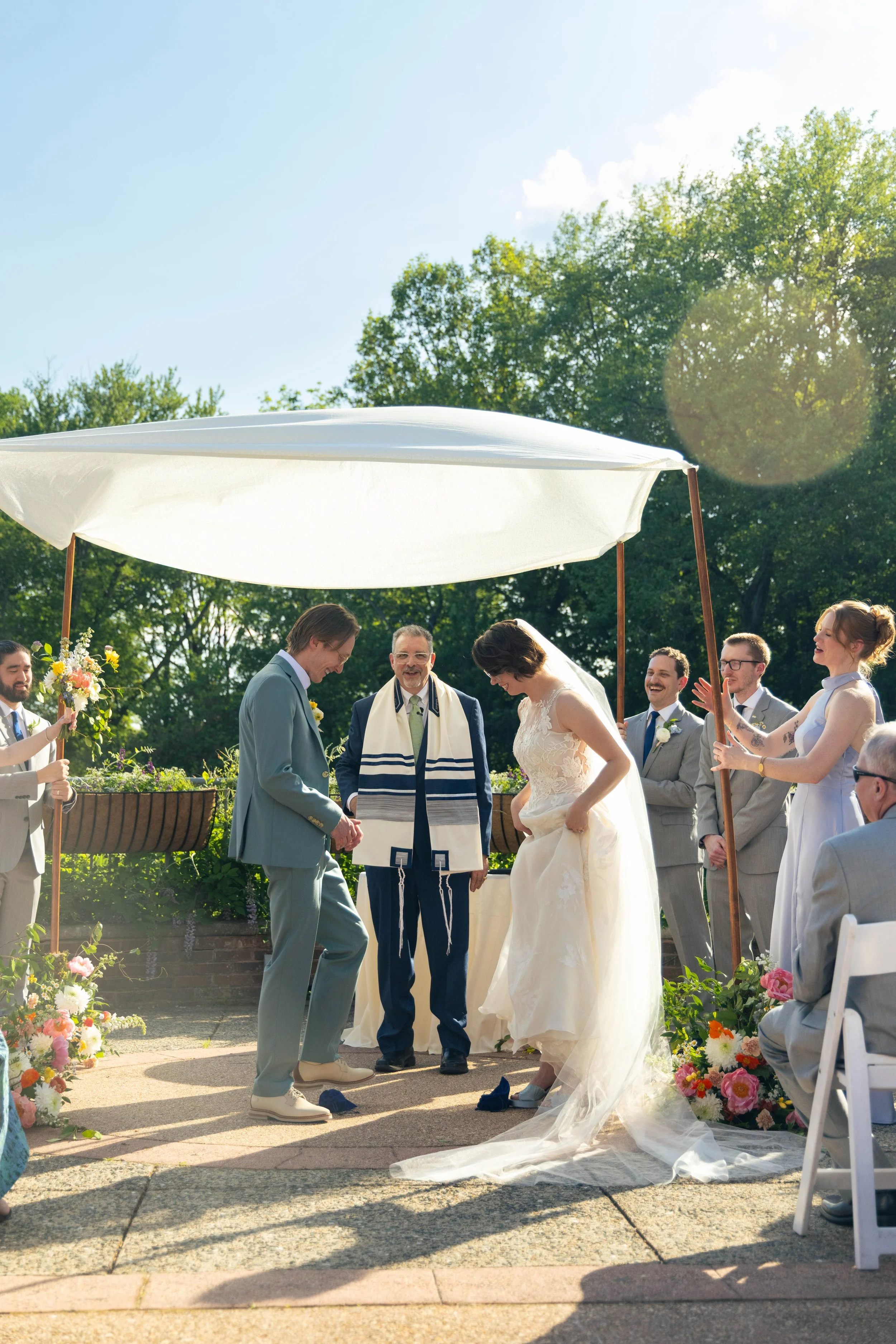 A wedding ceremony outdoors with the bride and groom holding hands. An officiant stands between them, and guests are watching and smiling. There are flowers and an umbrella providing shade, with green trees and a blue sky in the background.