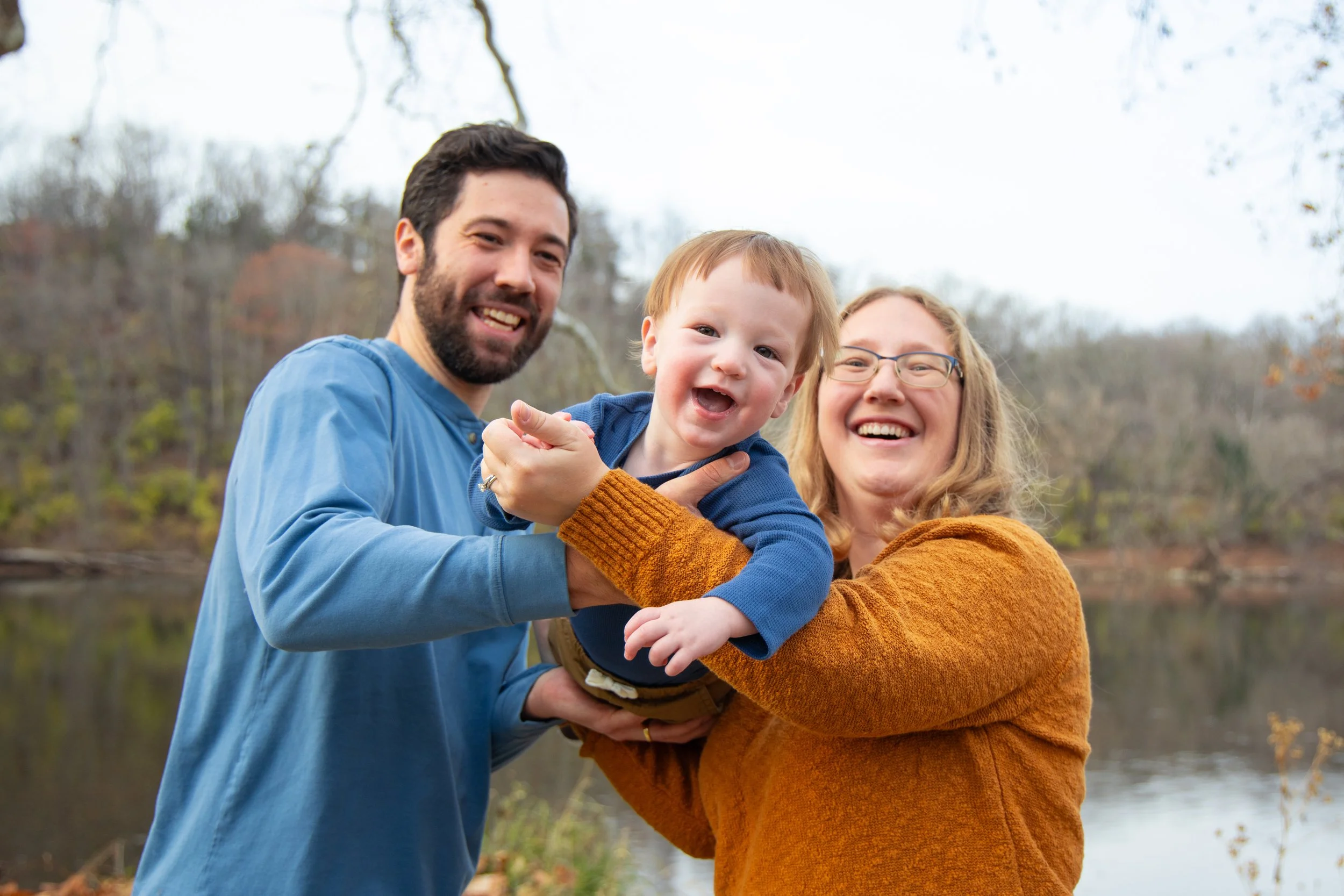 A family of three outdoors near a river, smiling and holding a young child, during autumn with trees in the background.